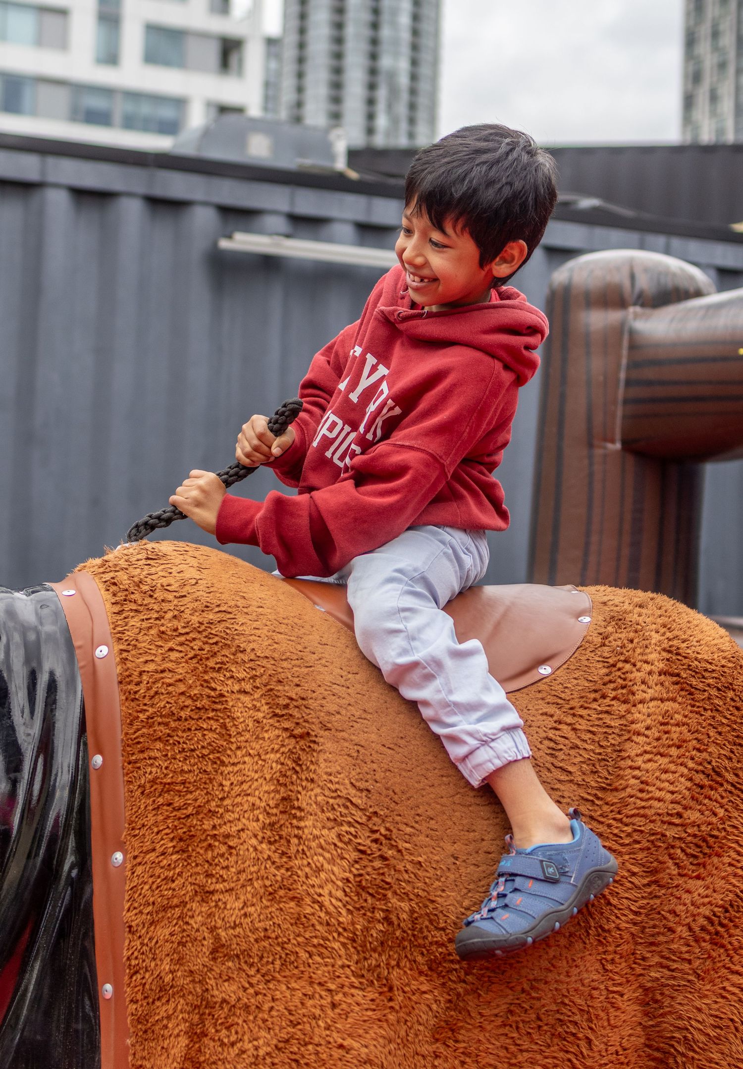 Smiling Asian boy in red hoodie riding a mechanical bull at an outdoor playground.