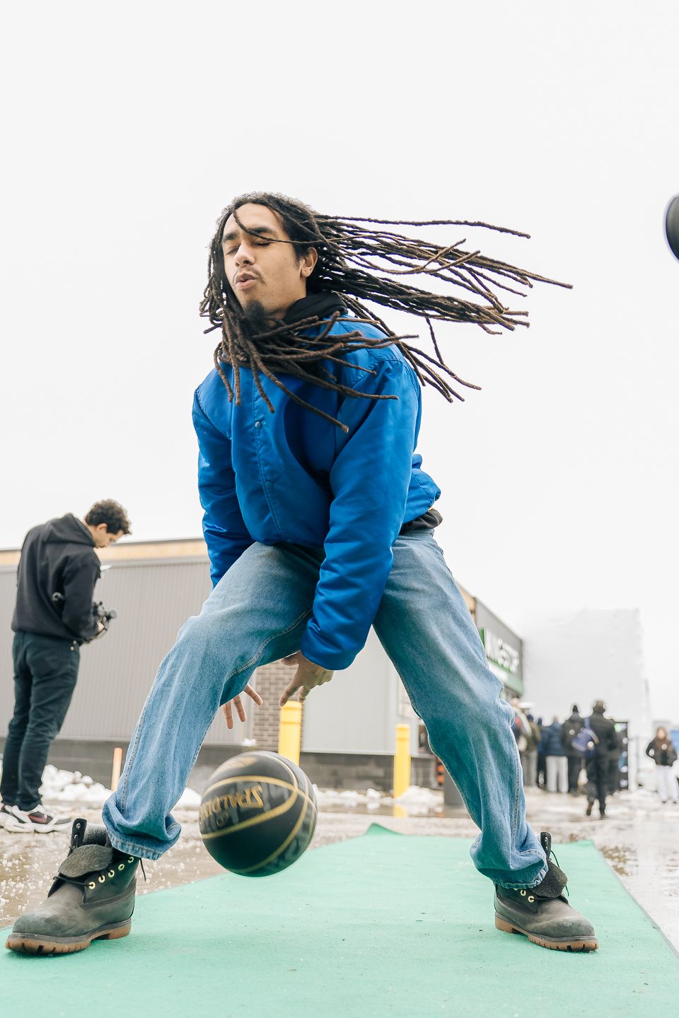 Young man with long dreadlocks dribbling basketball outdoors in blue hoodie and jeans on snowy day.
