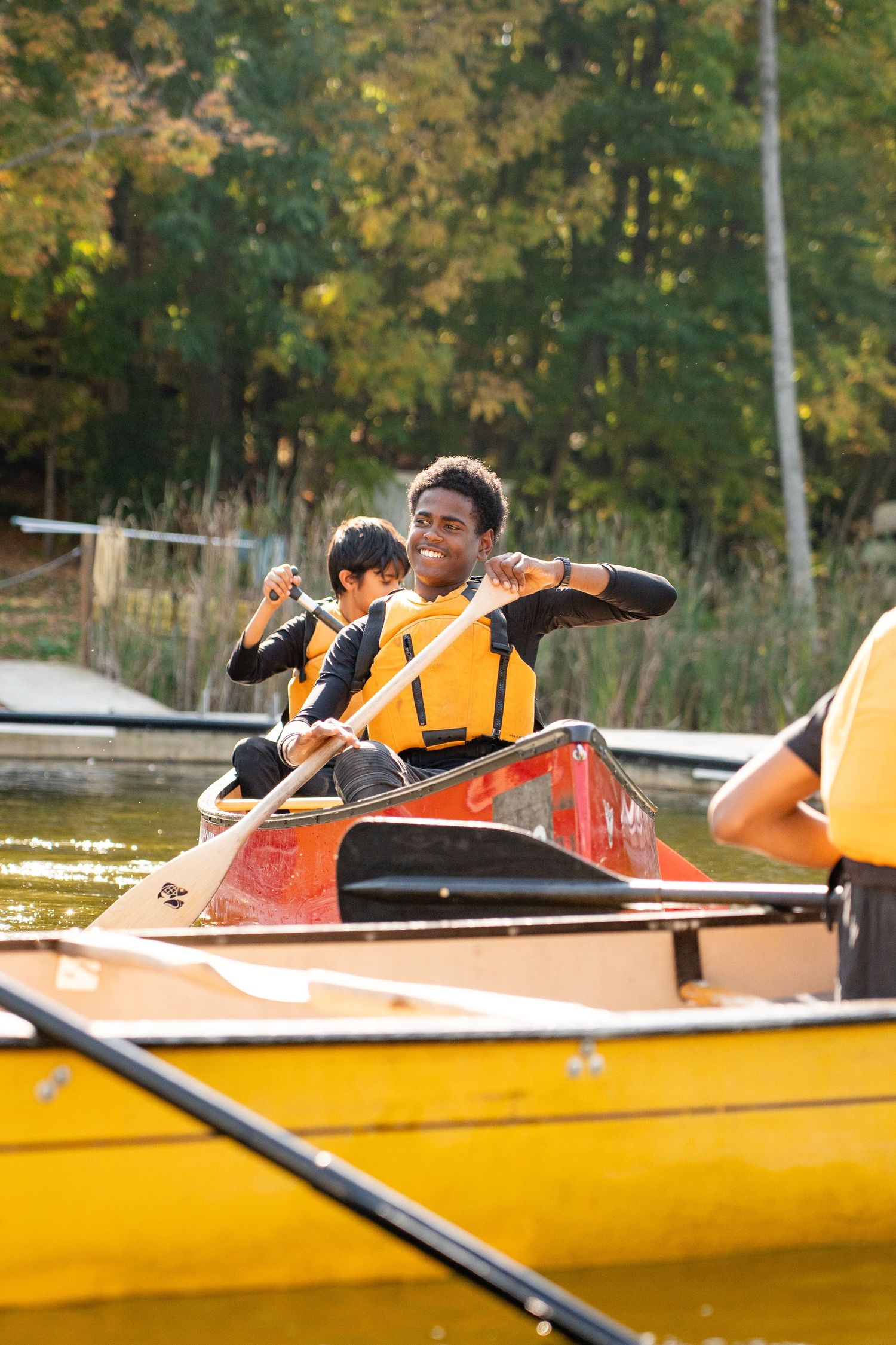 Happy young man paddling a red canoe wearing an orange life jacket on a calm lake.