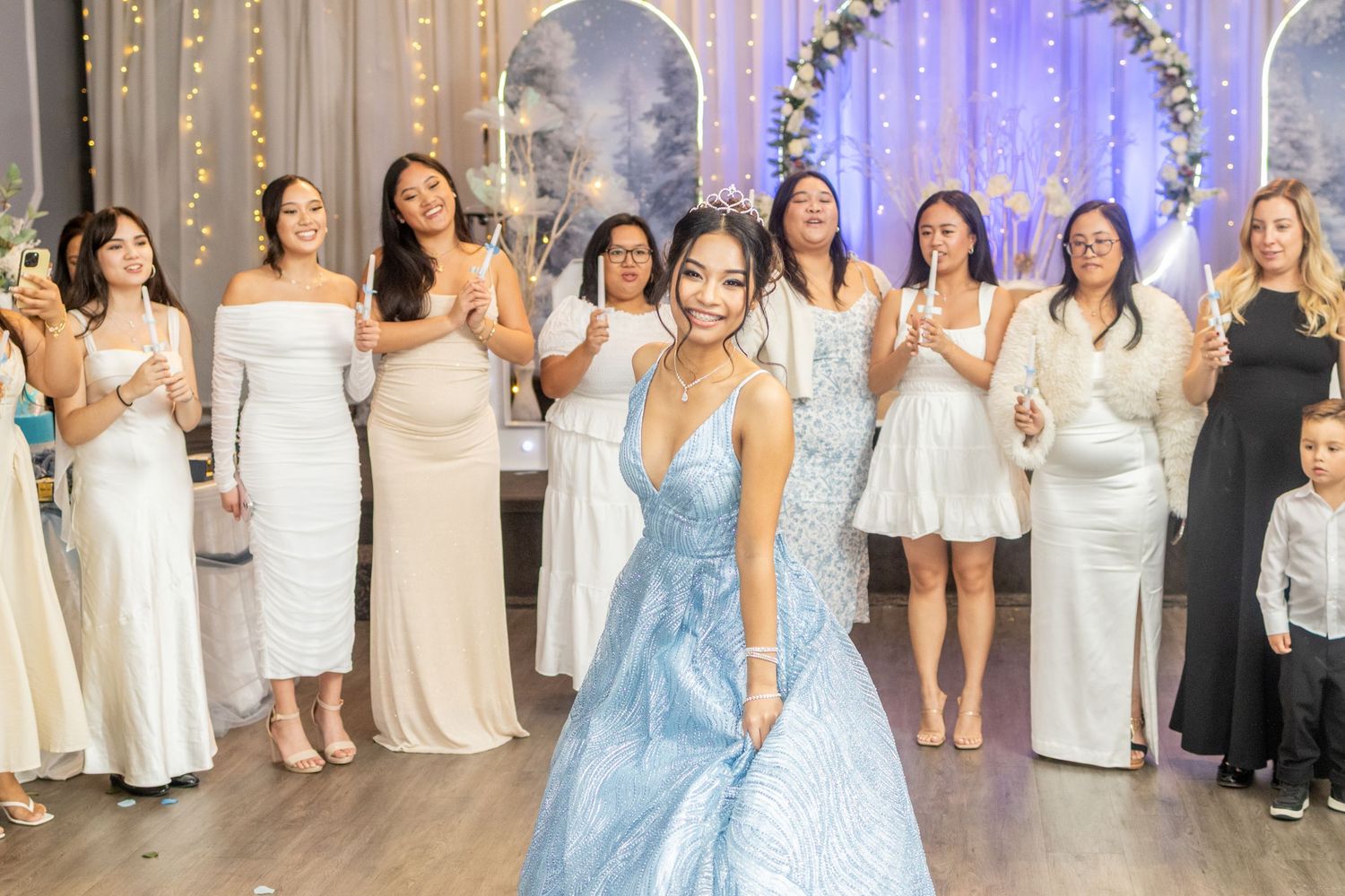 Teen girl in blue ball gown poses at quinceañera party with group of women in white dresses behind her.