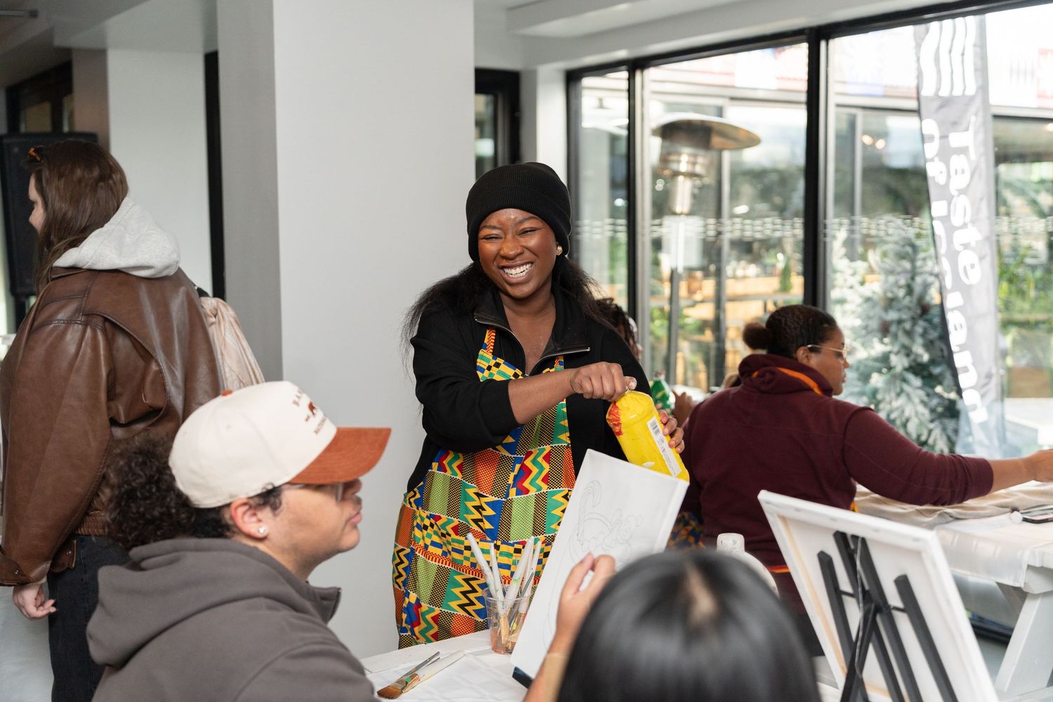 Smiling woman in colorful kente cloth apron teaching an art class at a bright indoor creative workshop.