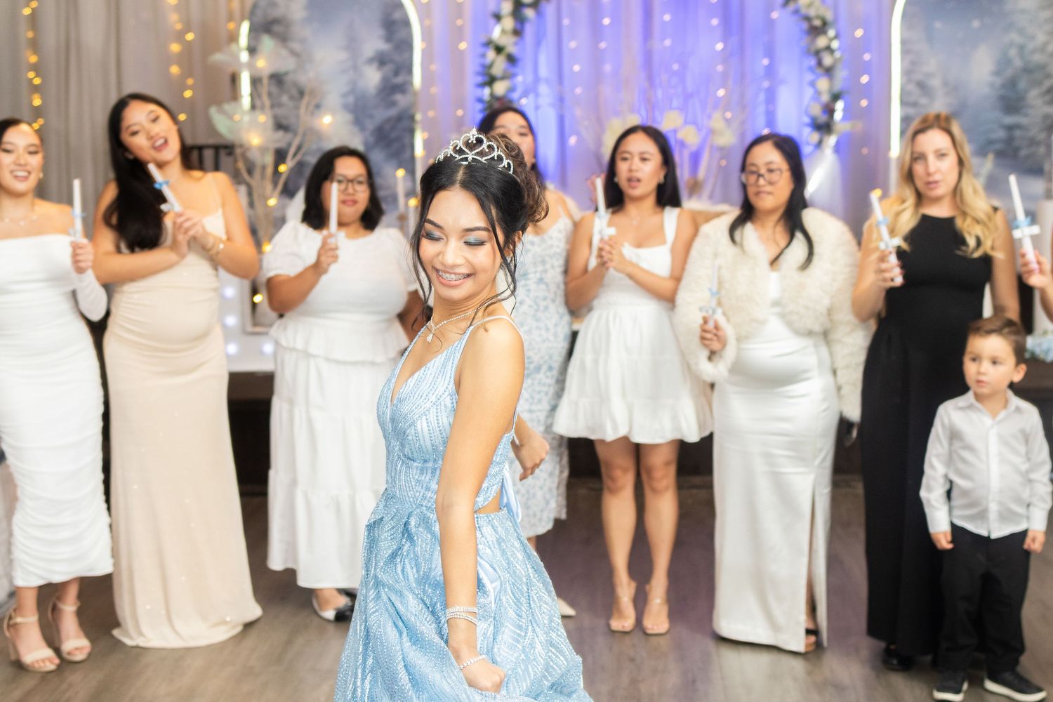 Young woman in blue ball gown dancing at quinceañera celebration with guests in white dresses holding candles.