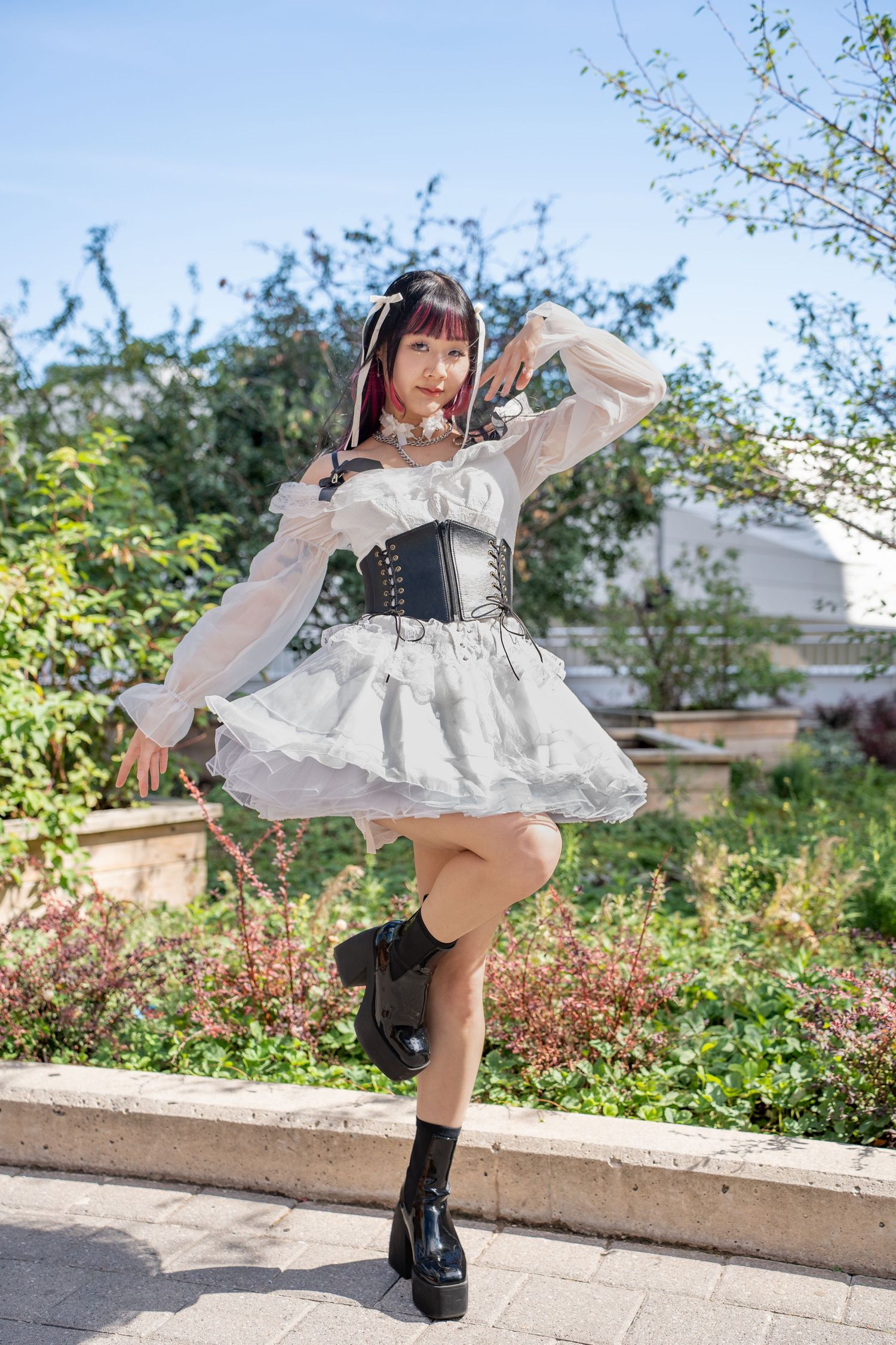 Young woman in white Lolita dress with black corset posing outdoors in a sunny garden.
