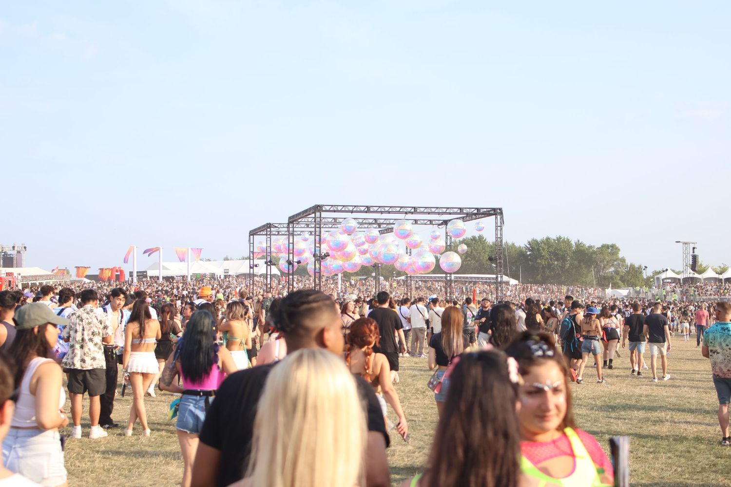Large crowd gathered at an outdoor music festival with a stage and balloons visible in the background.