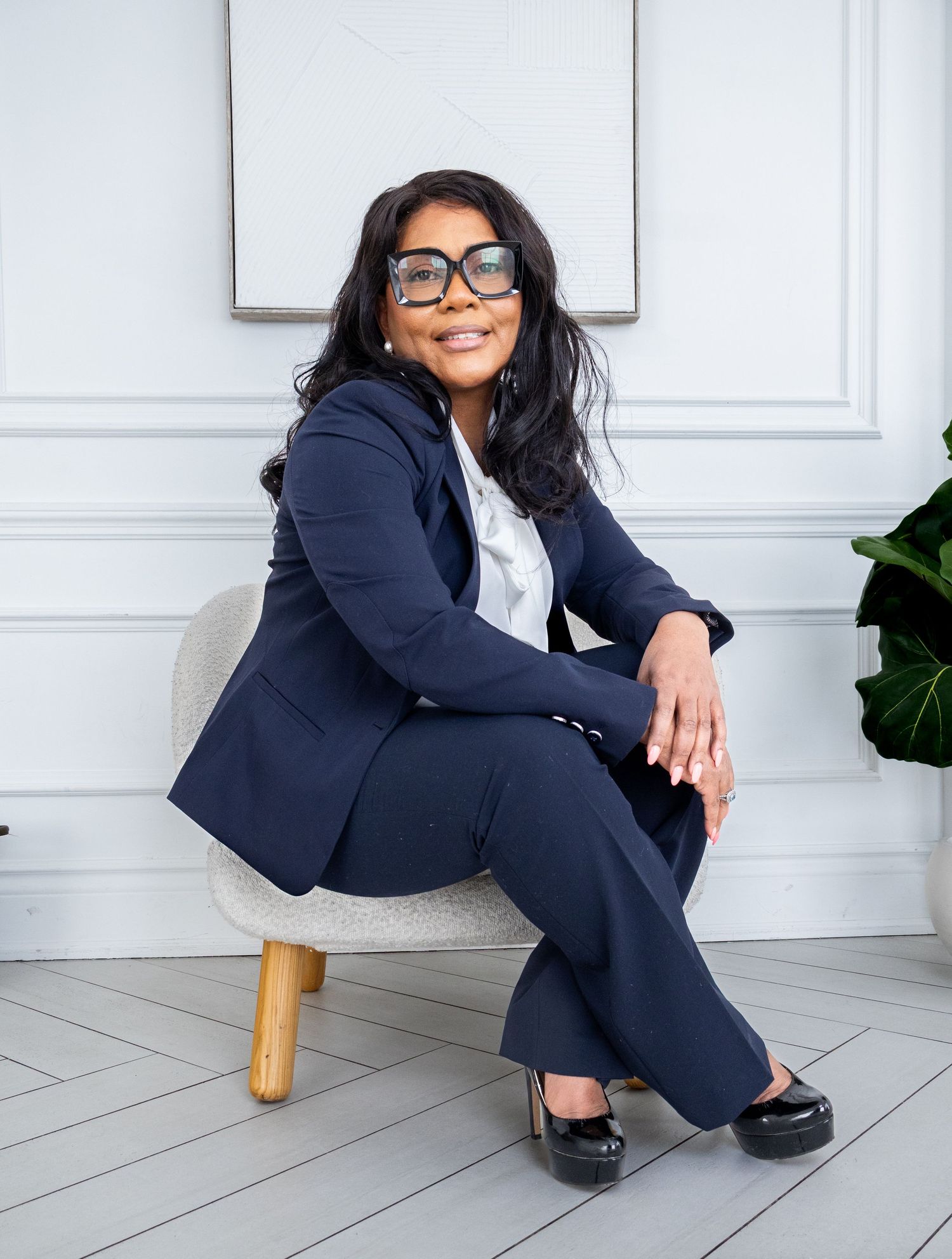 Professional woman in navy blue suit sitting confidently on a modern chair in a bright, elegant white interior.