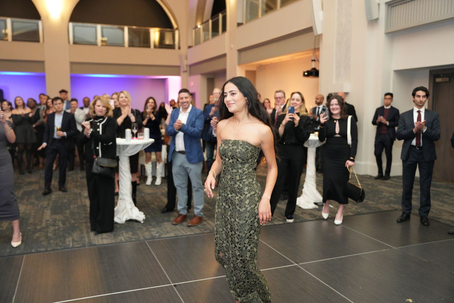 Woman in sequined dress walks across dance floor as guests mingle at an upscale evening event.