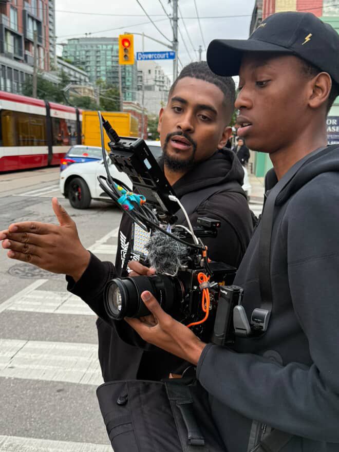 Two Black men reviewing camera footage on a busy urban street with a tram in background.