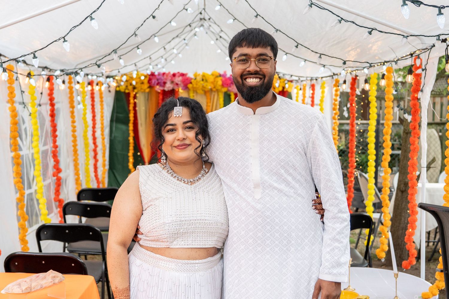 Couple in white outfits smiling together under string lights and floral garlands at Haldi ceremony.