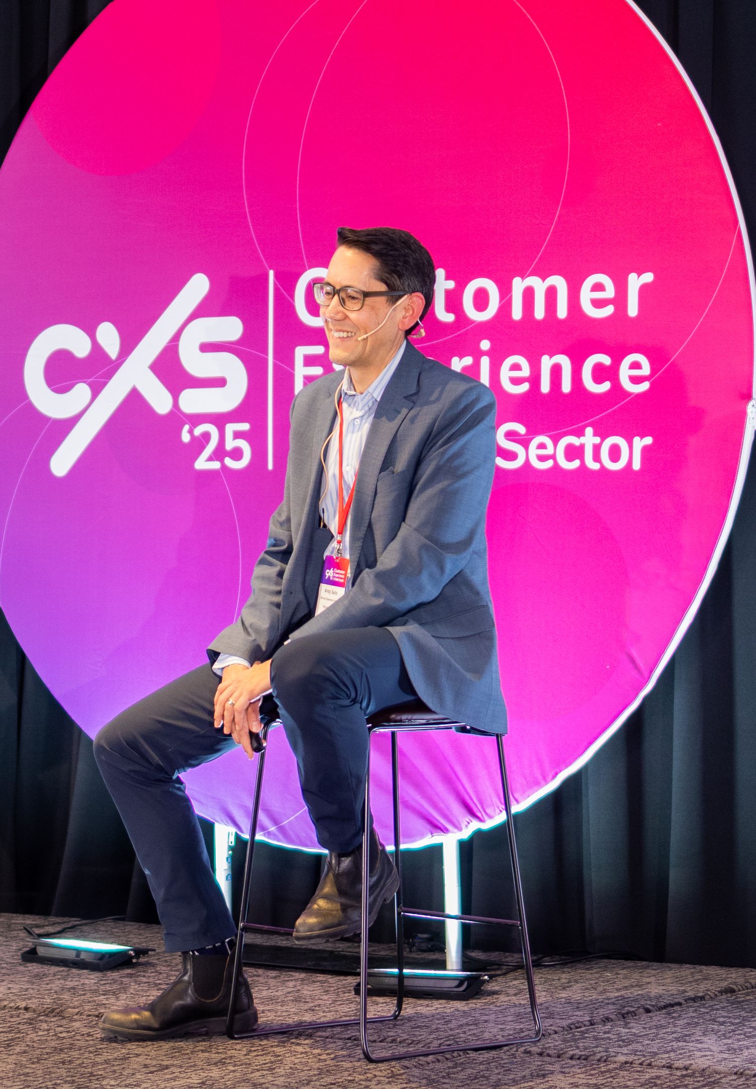 Male speaker sits on stool presenting at CXS 2025 Customer Experience for the Public Sector conference stage.