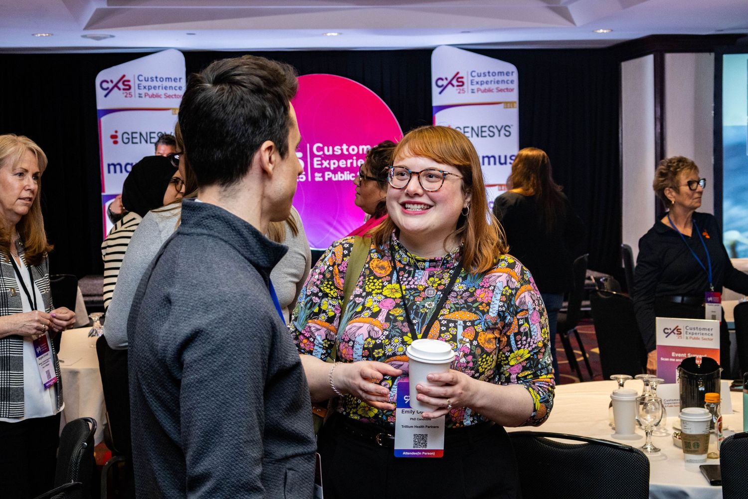 Woman in floral top holding coffee cup laughs while networking at CXS Customer Experience conference.