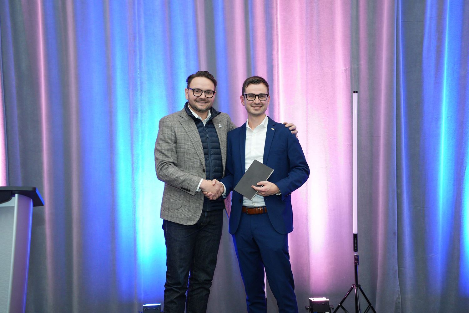 Two men shaking hands on stage at a corporate awards ceremony with colorful backdrop.