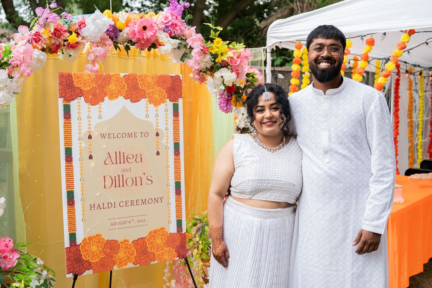 Couple posing beside Alliea and Dillon Haldi ceremony welcome sign with floral decorations.
