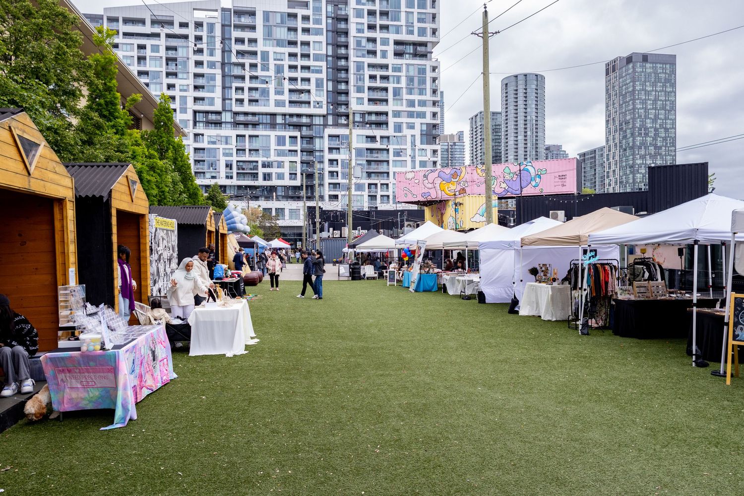 Outdoor vendor market with white tents and small wooden booths on green turf with city skyline backdrop.