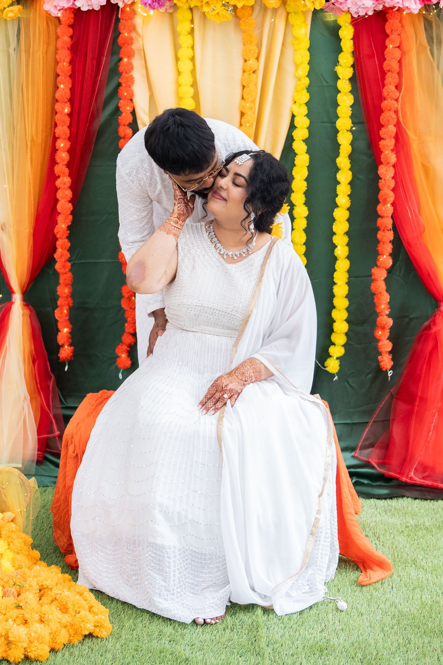 Groom tenderly kissing bride at colorful Indian Haldi ceremony with marigold garland backdrop.