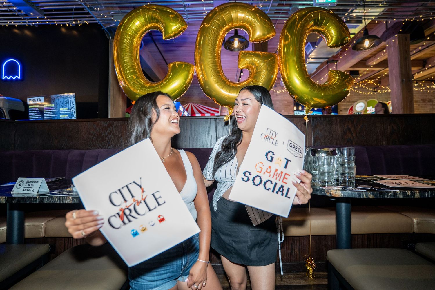Two women posing with City Circle Game Social signs under gold CGC balloons at venue.