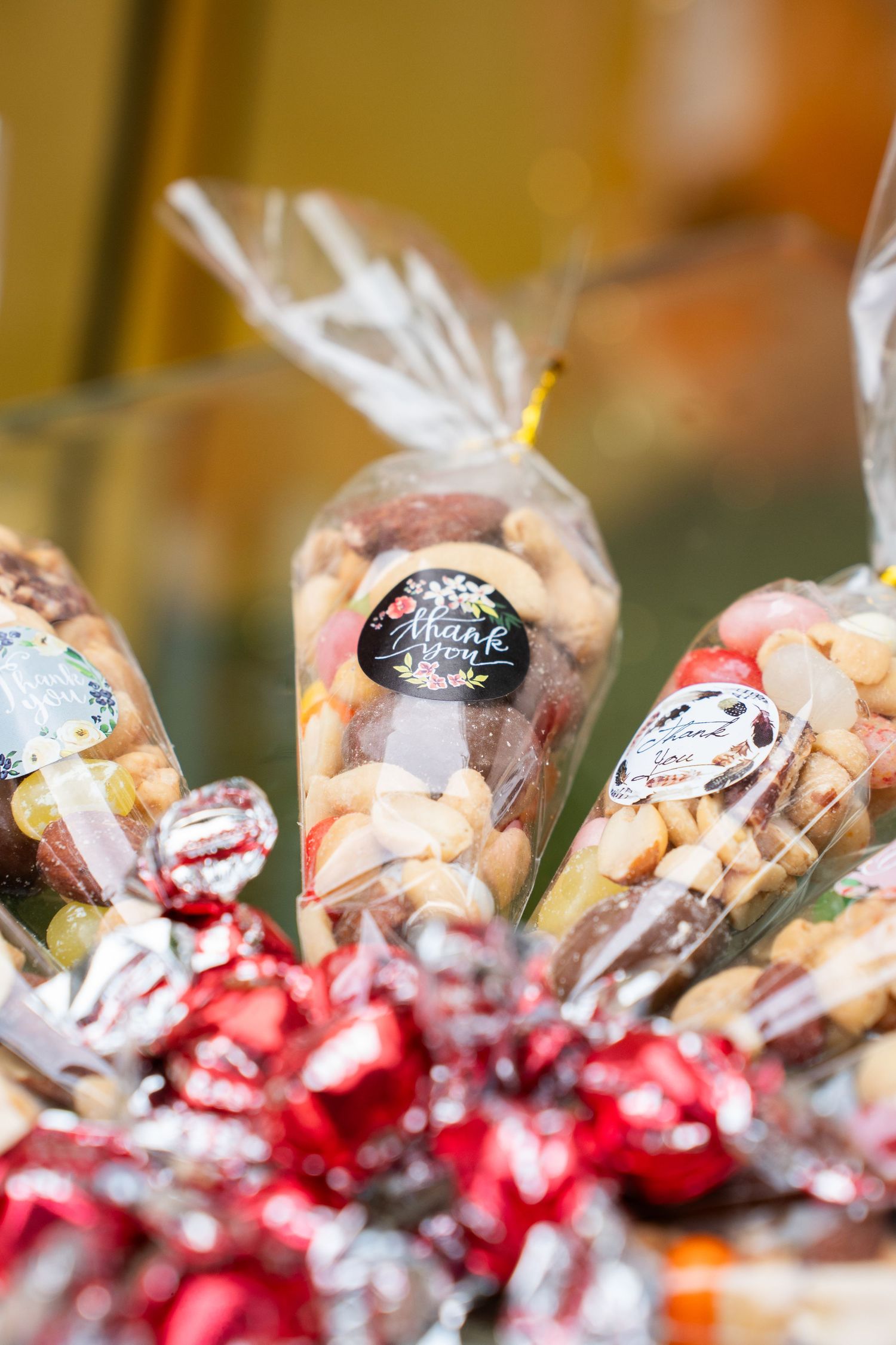 Transparent gift bags filled with mixed nuts and thank you stickers surrounded by red candies.