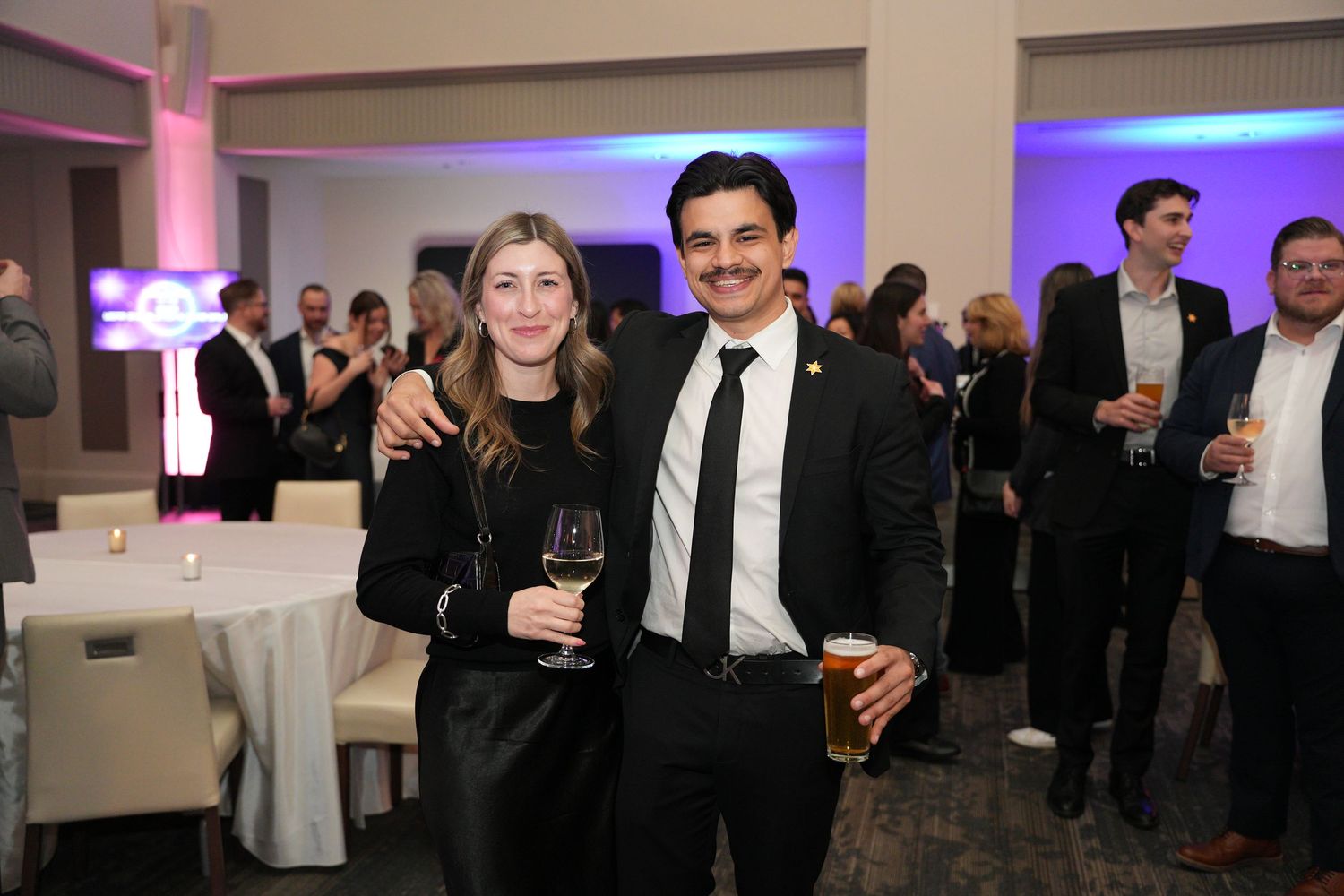 Couple smiling and holding drinks at an elegant evening corporate event with purple lighting.