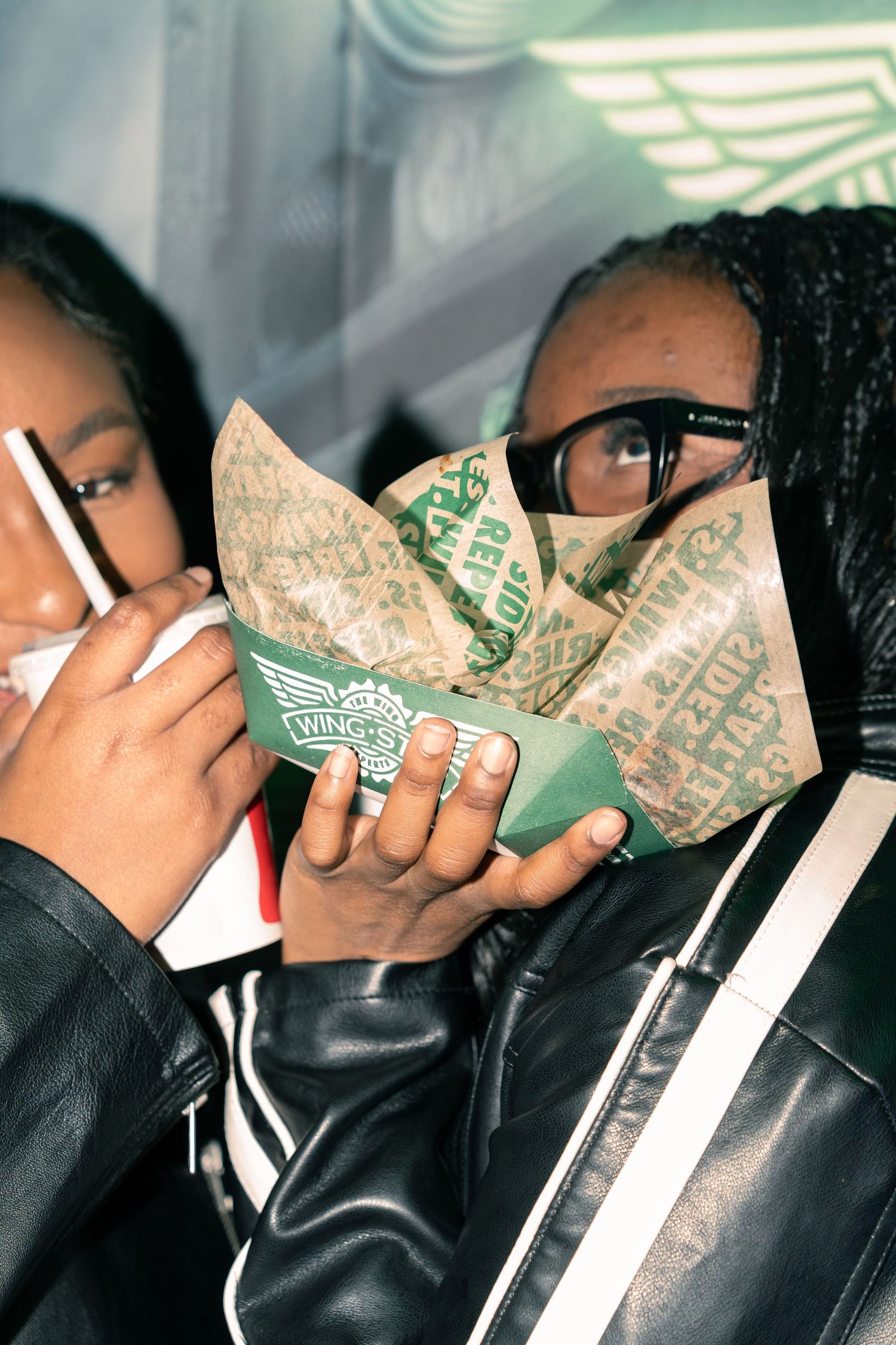 Person in leather jacket holding Wings restaurant branded chicken wing box close to face.