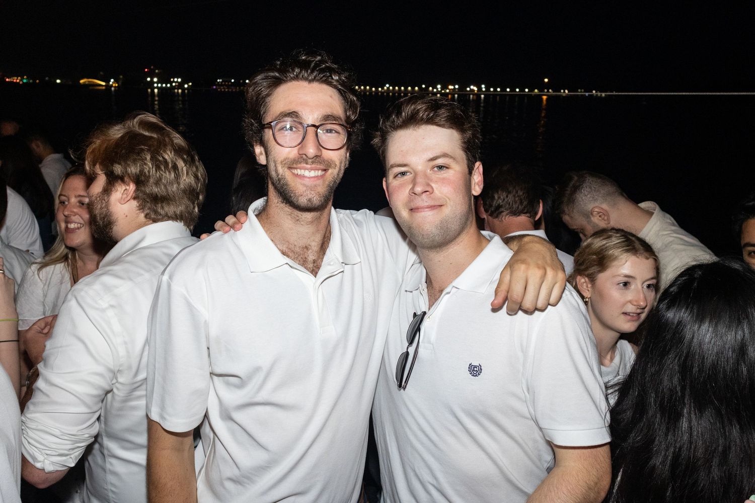 Two smiling men in white shirts posing together at a nighttime waterfront party.