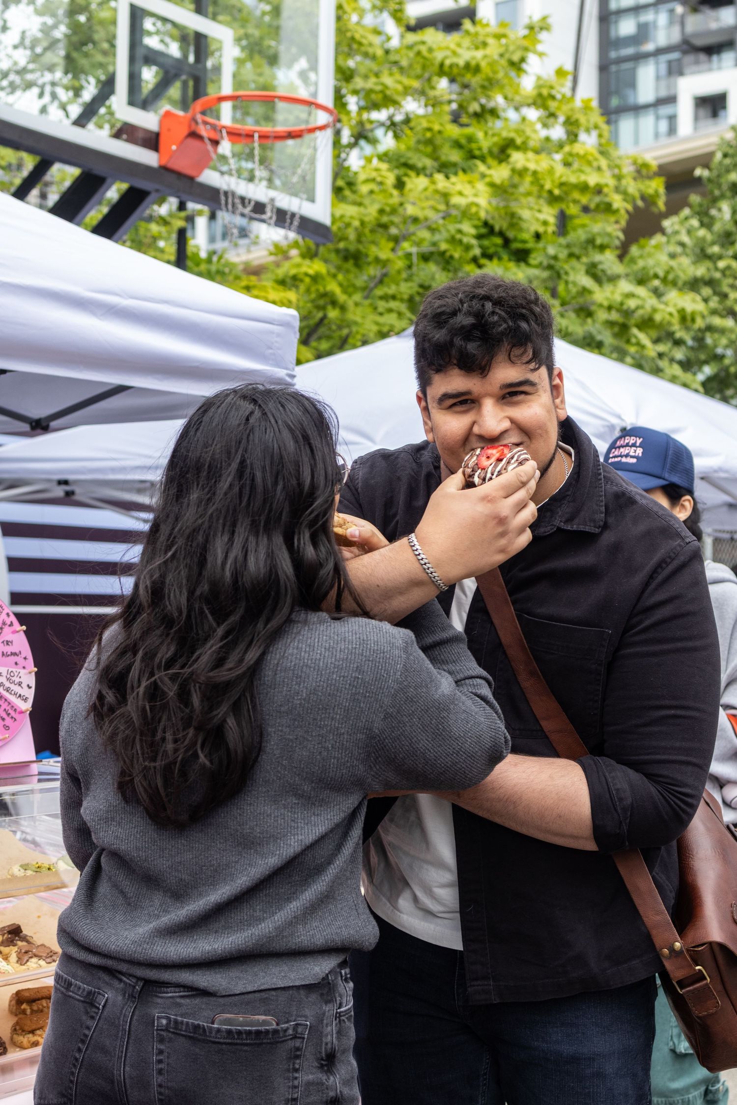 Woman feeding a chocolate donut to smiling man at outdoor food market festival event.