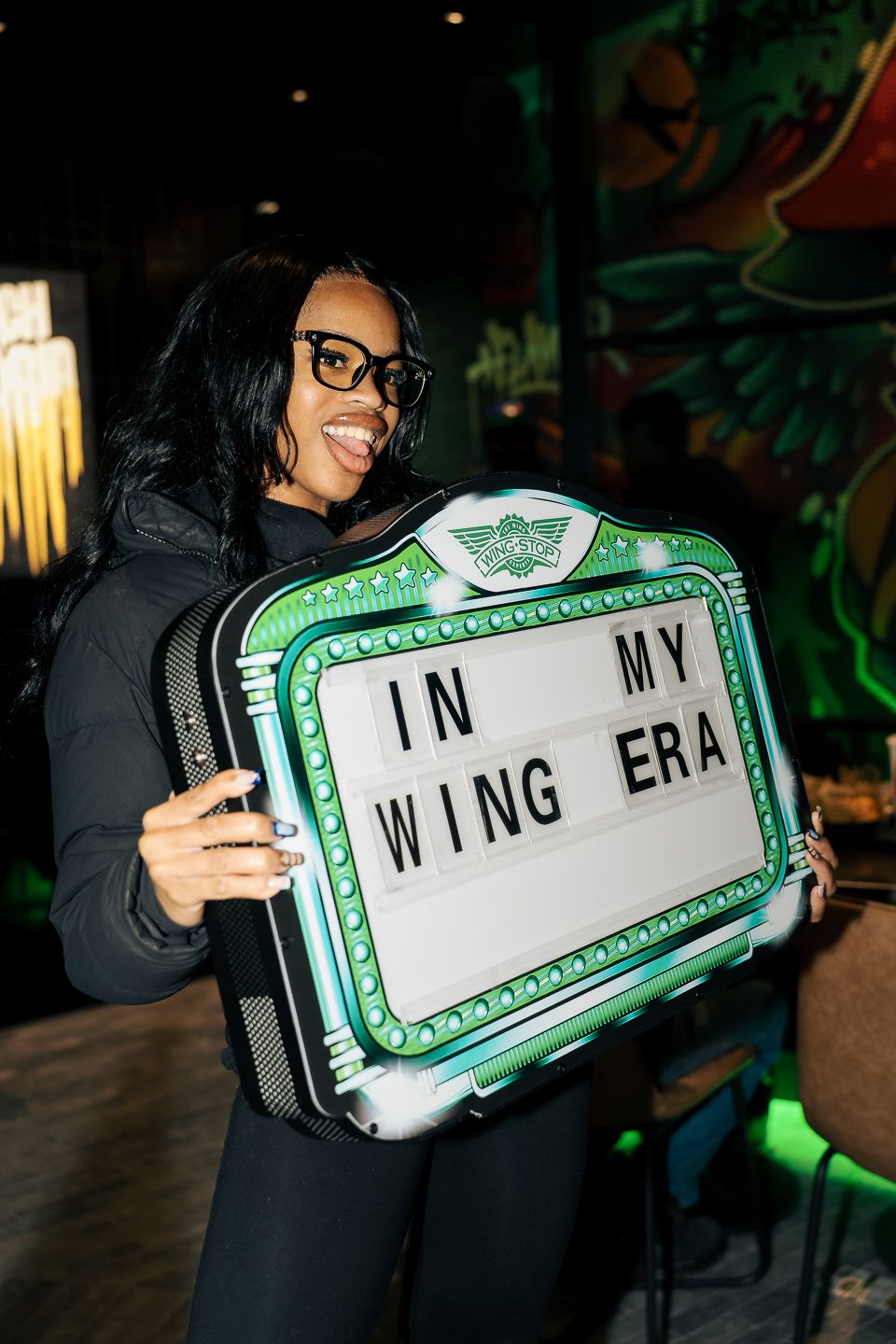 Woman in black outfit holding illuminated 'In My Wing Era' sign at a restaurant.