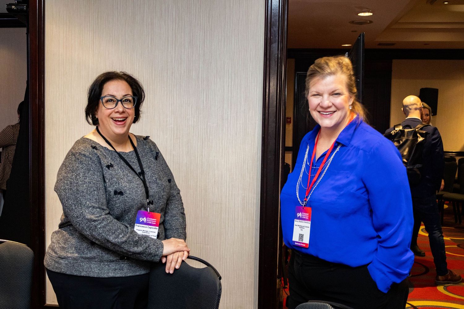 Two professional women wearing conference lanyards smiling together at CXS public sector business event.