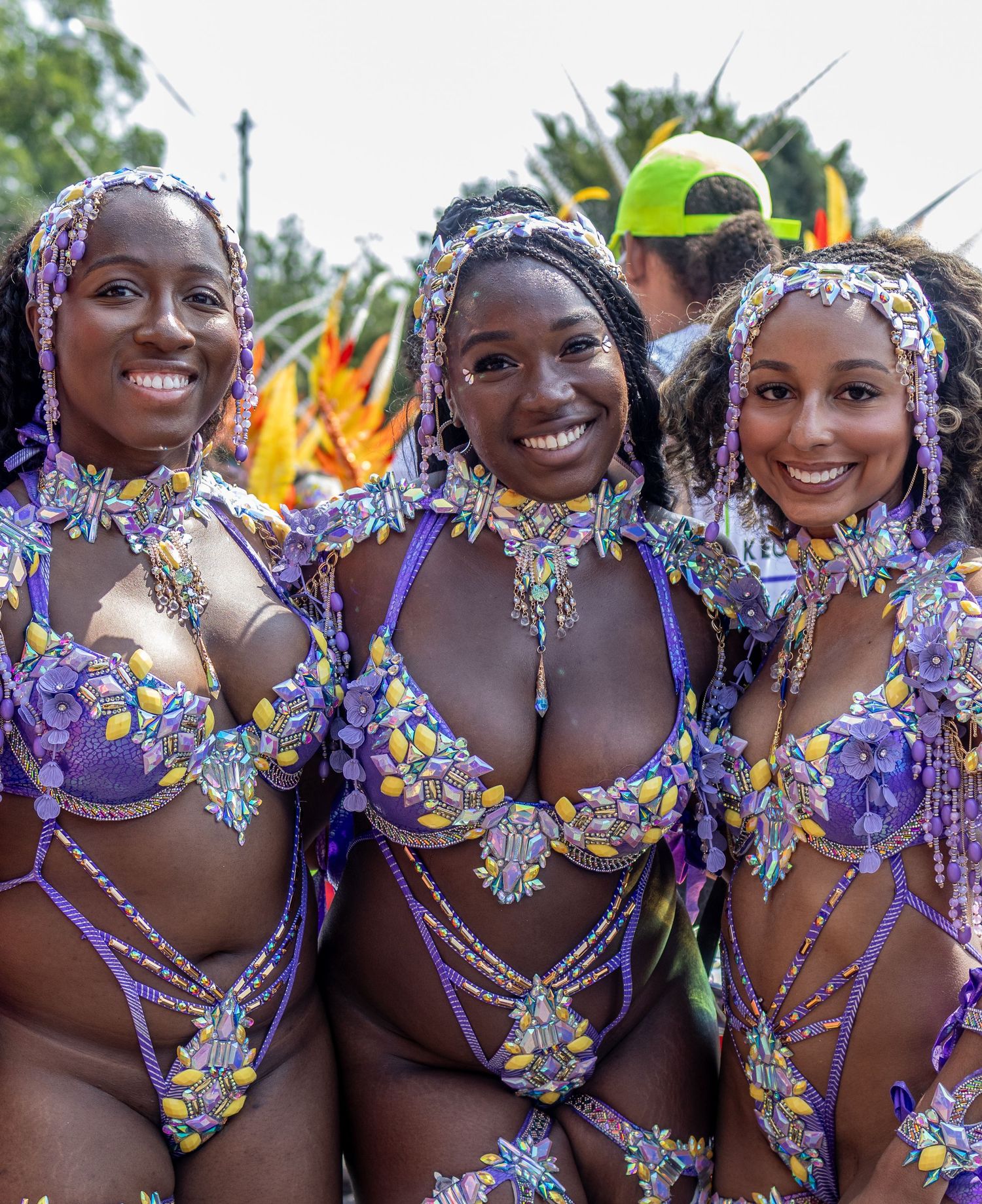 Three smiling women in matching purple beaded carnival bikini costumes at a street festival parade.