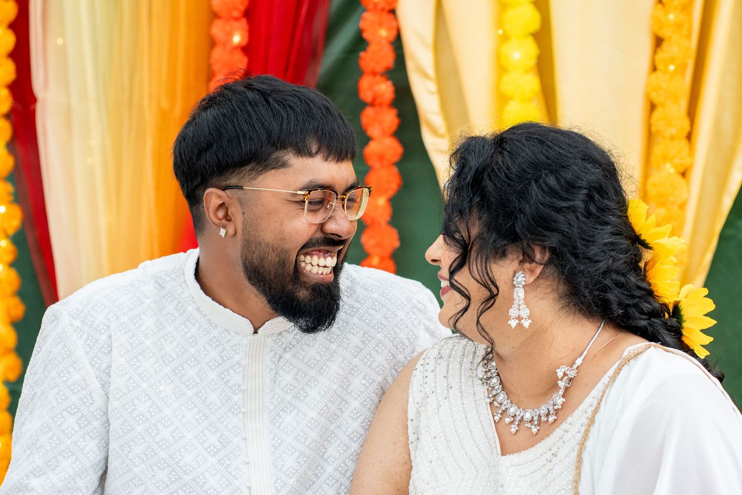 Laughing couple in white traditional Indian attire at a colorful Haldi ceremony celebration.