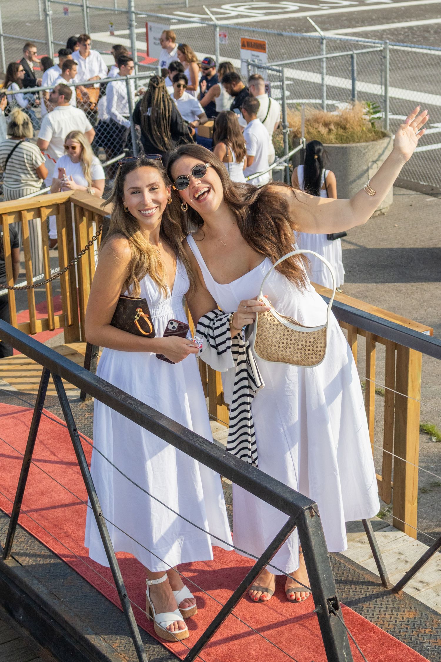 Two women in white dresses posing on red carpet stairs at an outdoor event.
