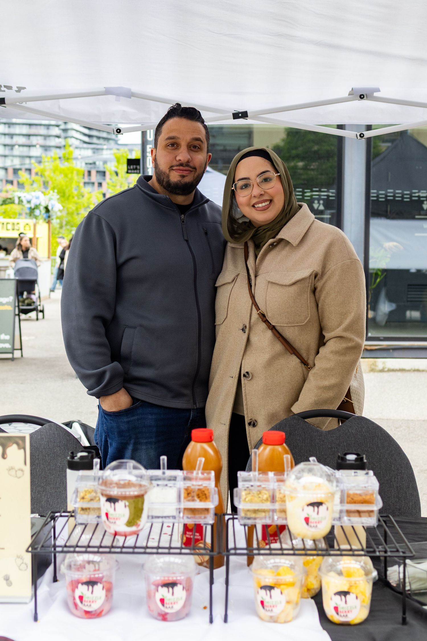 Couple smiling behind a food stall with colorful bottled drinks and packaged snacks at an outdoor market.