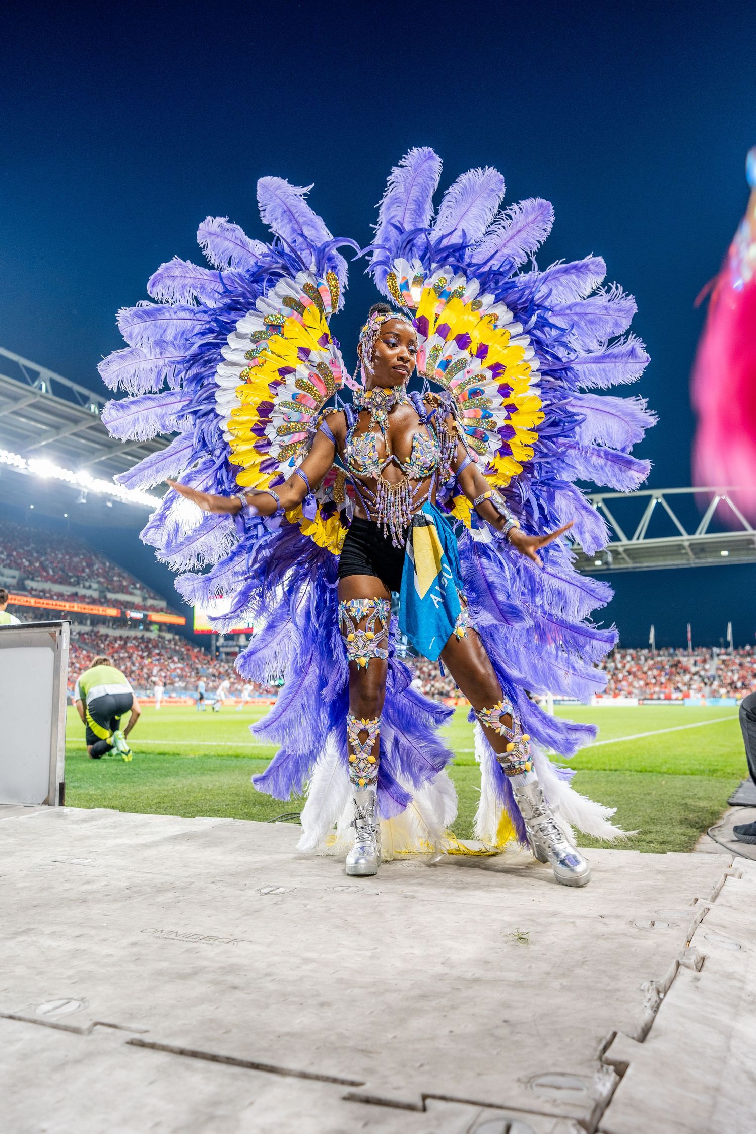 Performer in vibrant purple and yellow feathered carnival costume dancing at a stadium event.