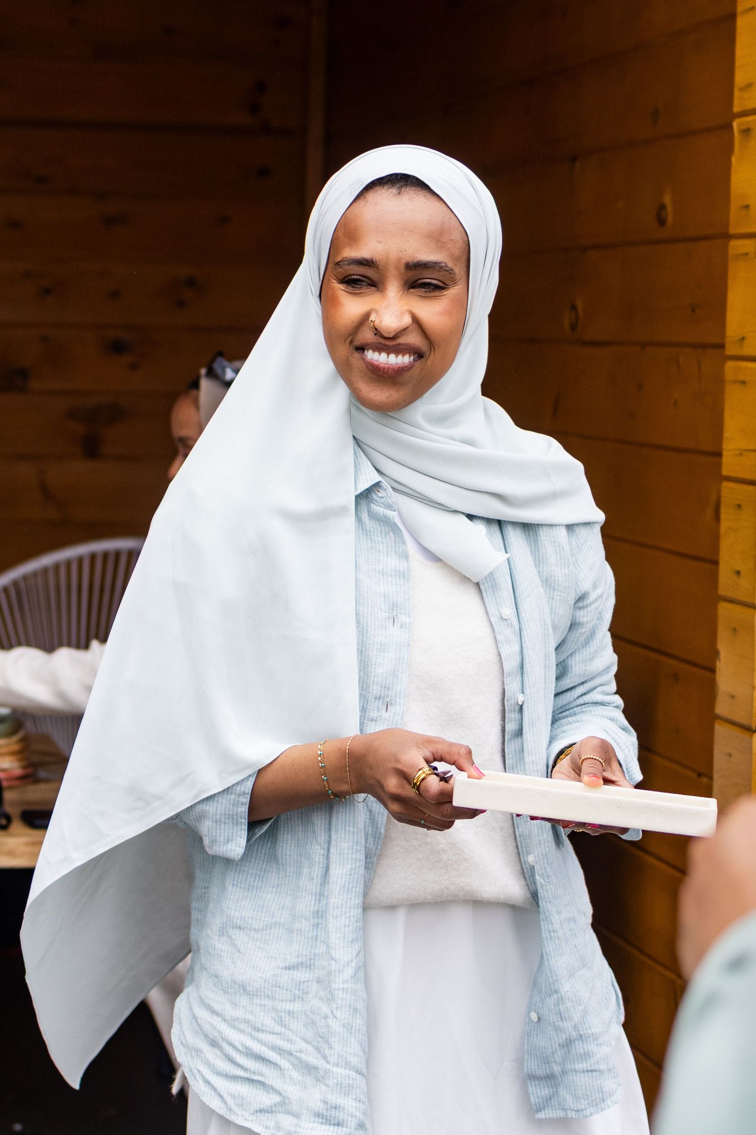 Smiling Black woman in white hijab and light blue cardigan holding a book outdoors.