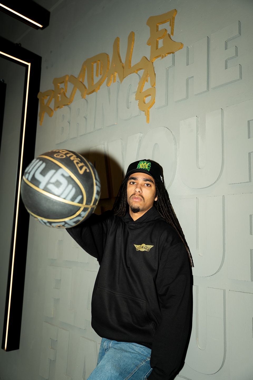 Man with dreadlocks holding basketball posing indoors beside gold Rexdale graffiti lettering on white wall.