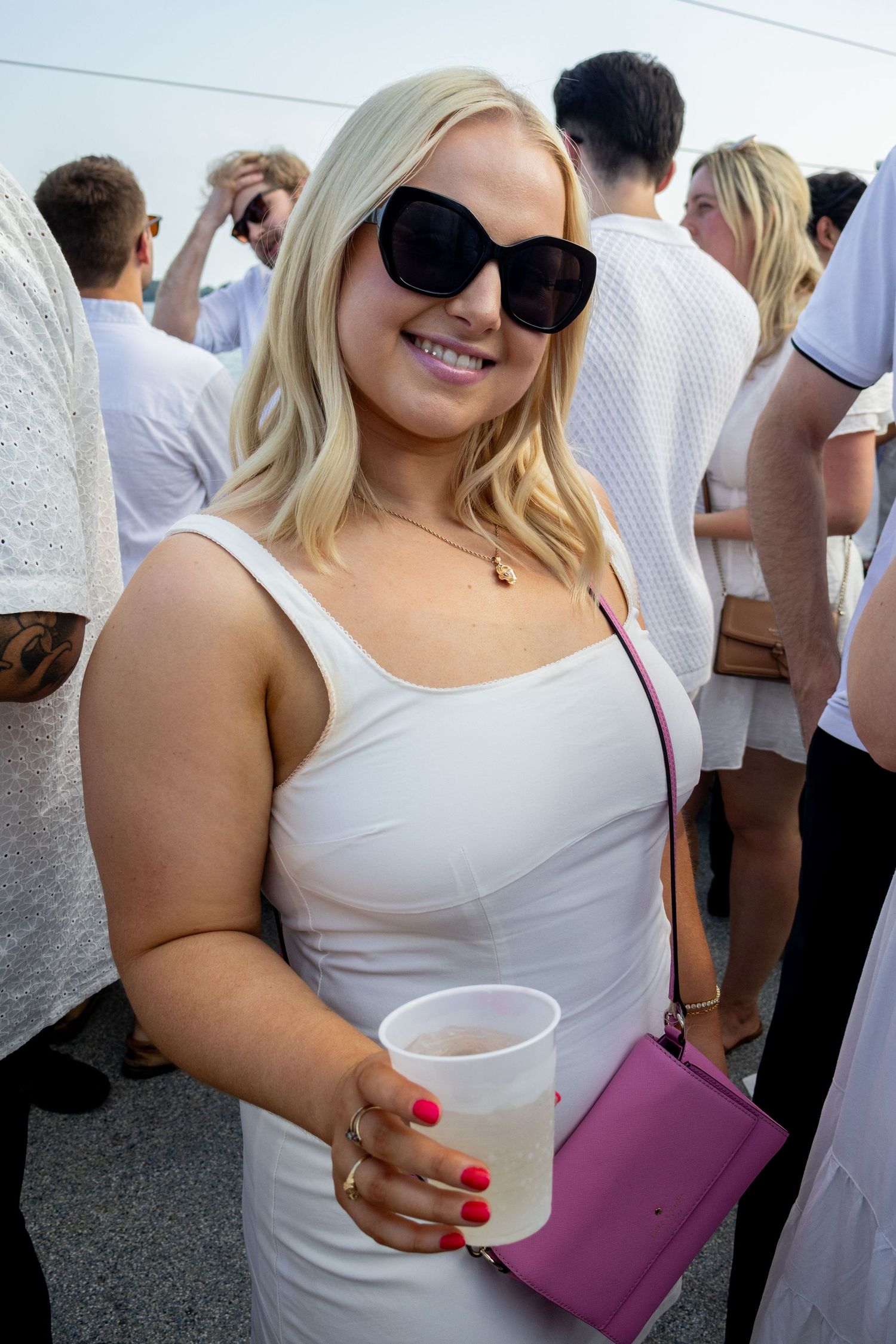 Smiling blonde woman in white dress holding a drink at an outdoor white party.