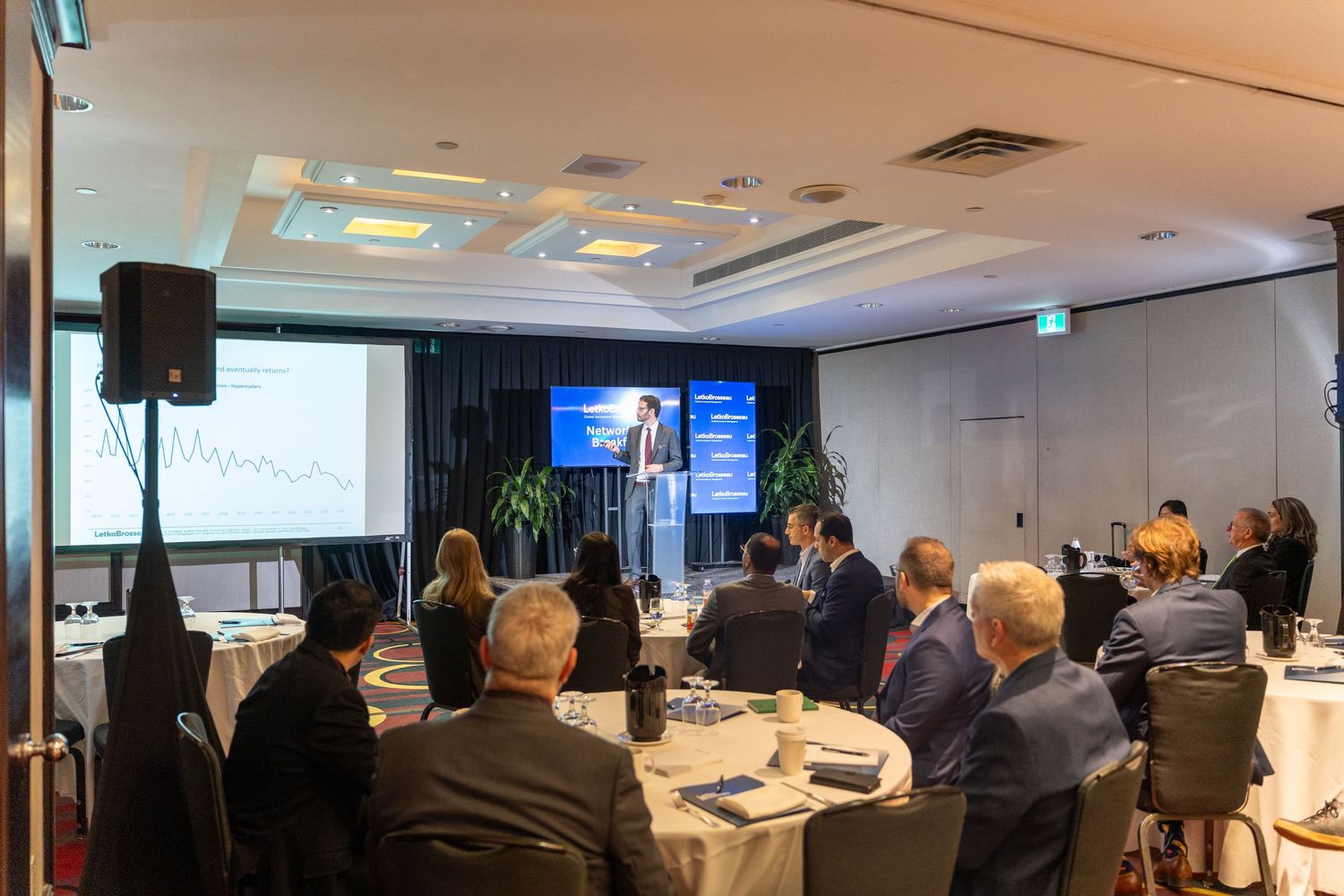 Business professionals attending a conference presentation with a speaker at a podium and projection screen.
