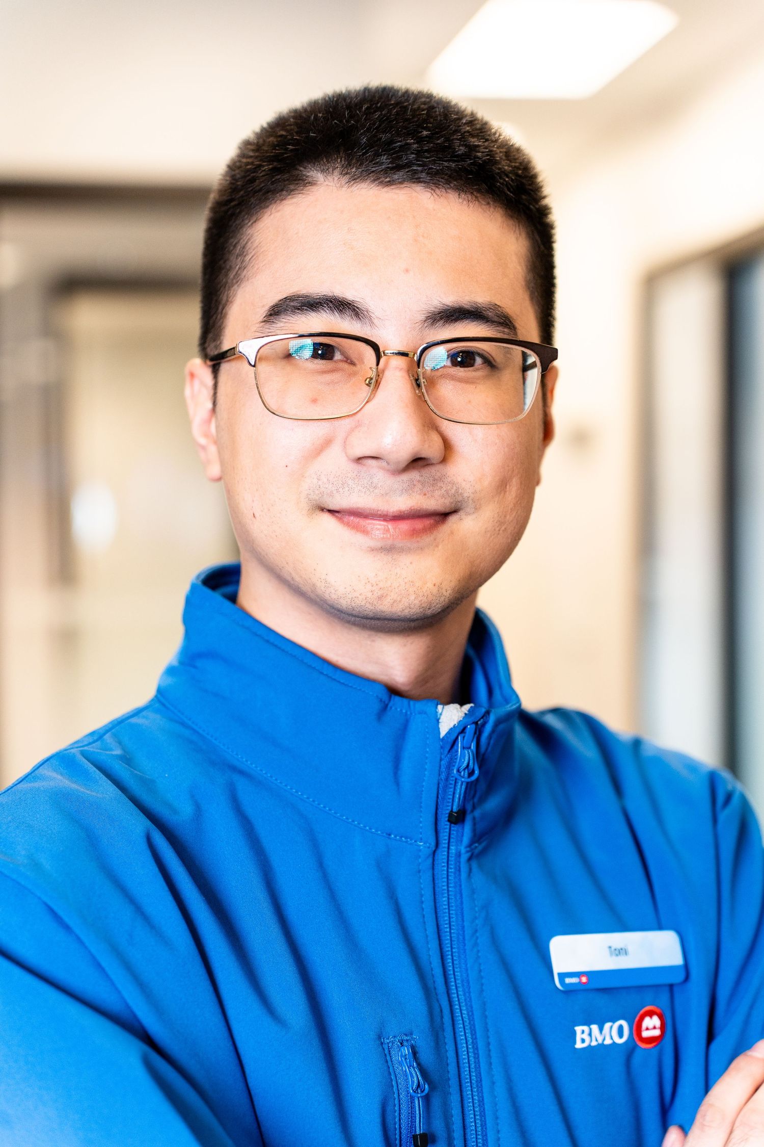BMO employee smiling in a blue branded jacket with glasses and arms crossed in an office hallway.