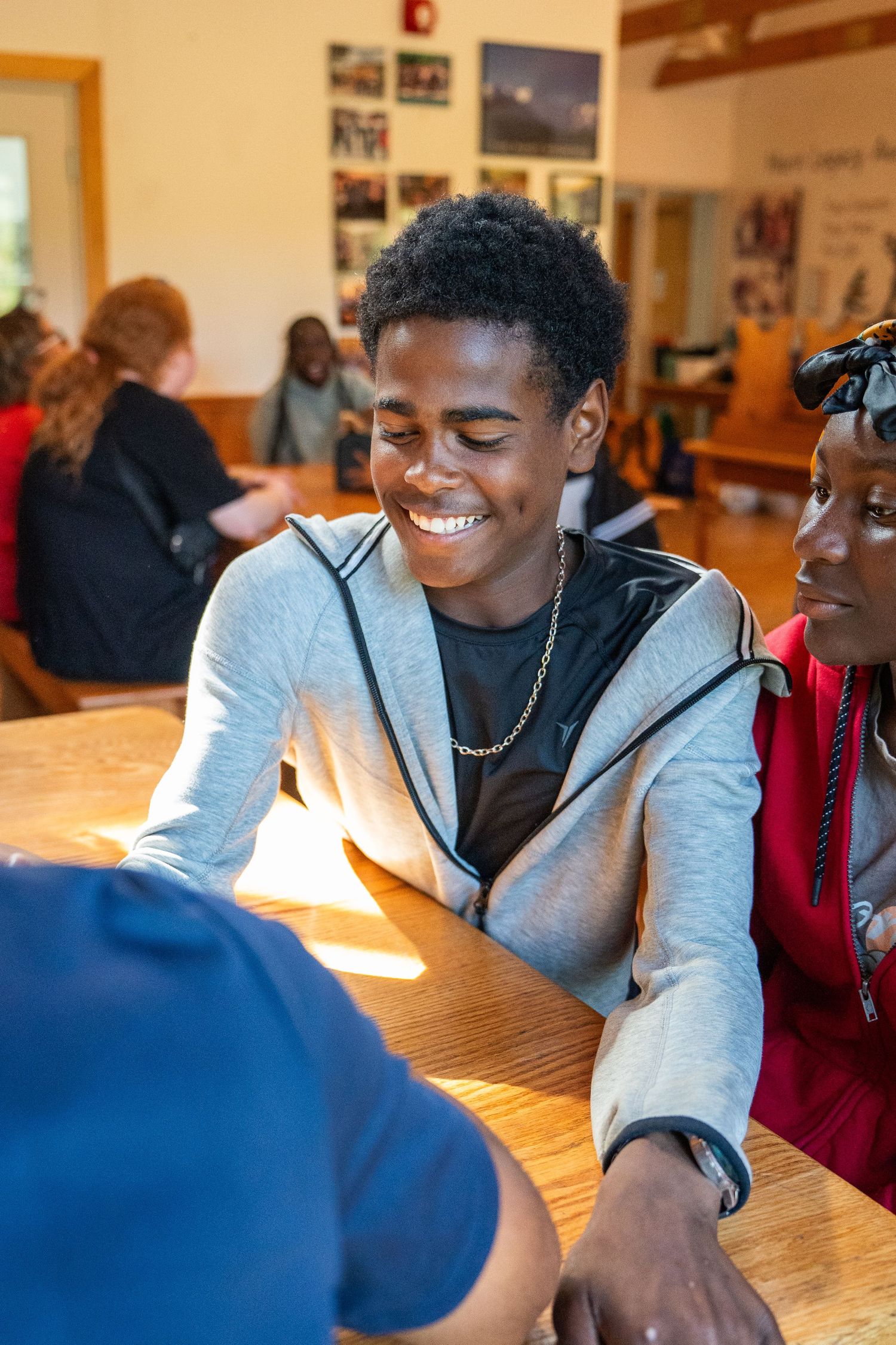 Smiling young man in gray hoodie enjoys conversation at a wooden table indoors.