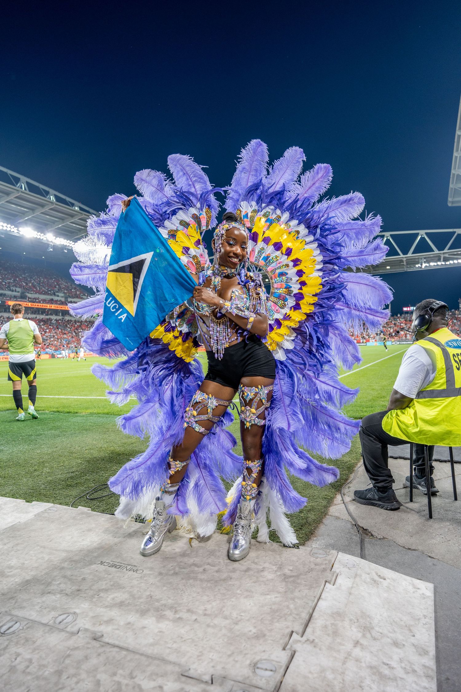 Performer in vibrant purple feathered carnival costume holding Saint Lucia flag at stadium event.