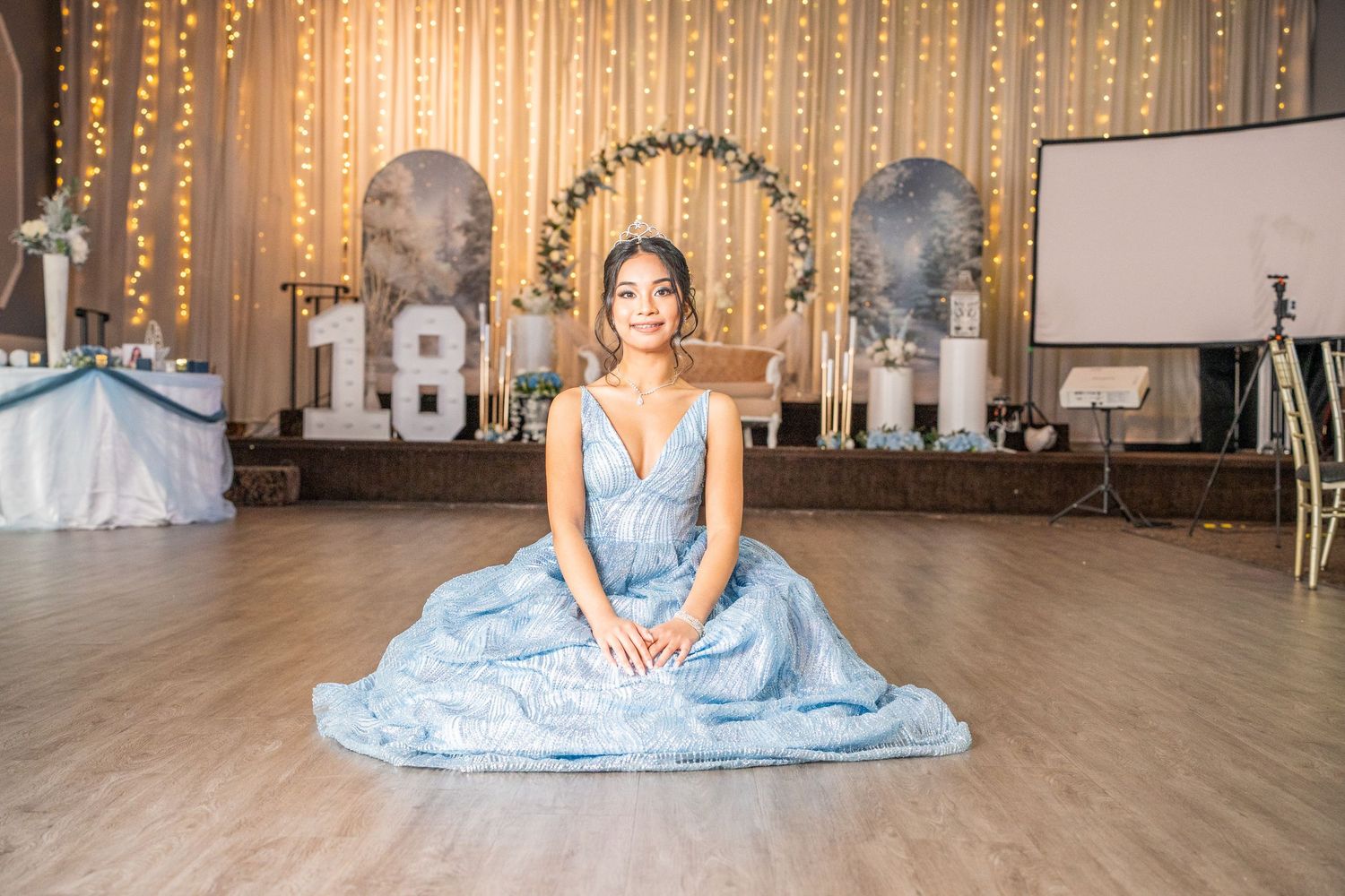 Young woman in light blue ball gown sits gracefully on floor in front of 18th birthday party decor.