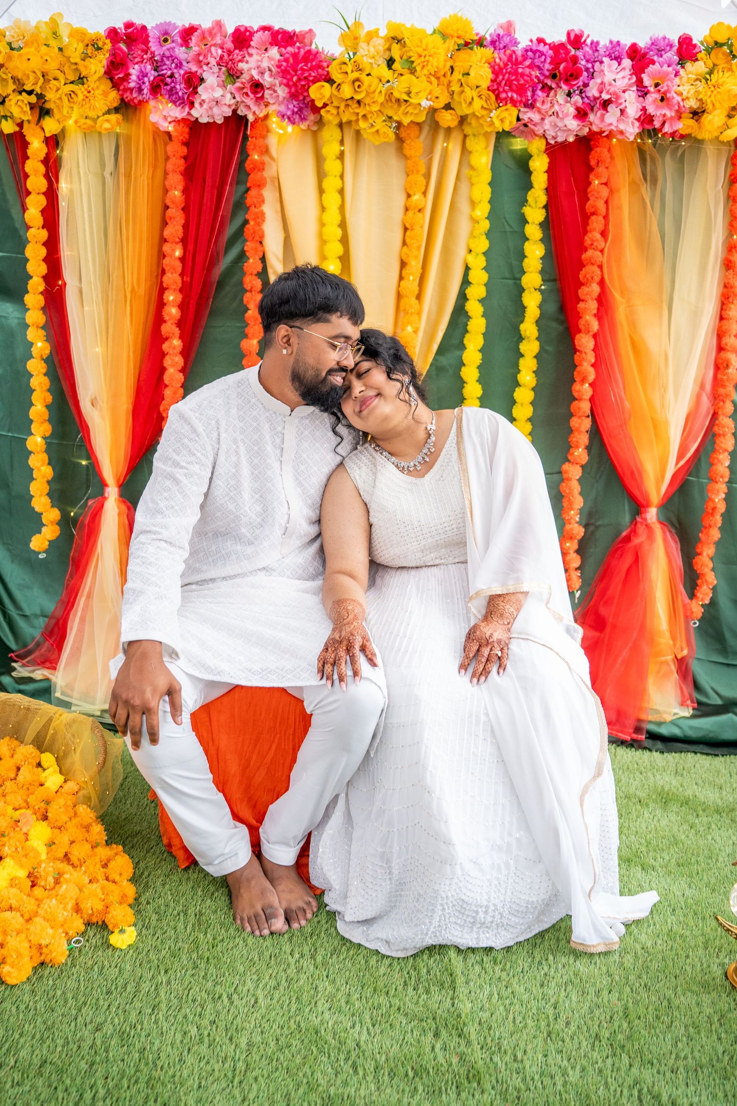 Couple in white outfits sits together under colorful marigold garlands at Indian celebration.