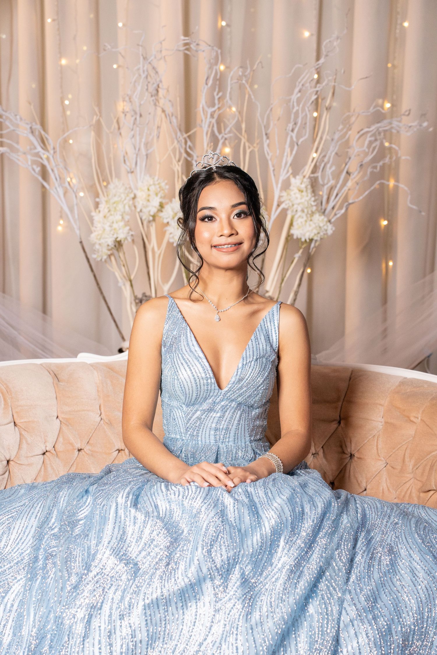 Teenage girl in sparkling light blue ball gown and tiara poses on elegant sofa at her debut party.
