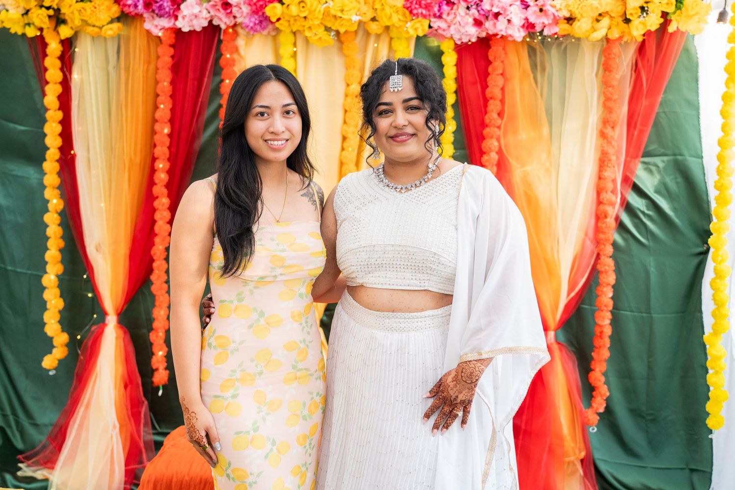 Two women in festive Indian attire pose together in front of colorful floral garland backdrop.