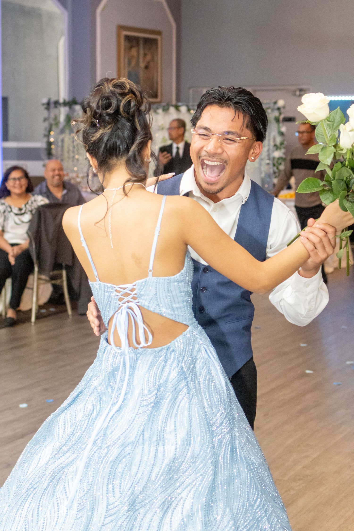 Young man in a vest joyfully dancing with a woman in a blue gown holding a white rose at a formal event.