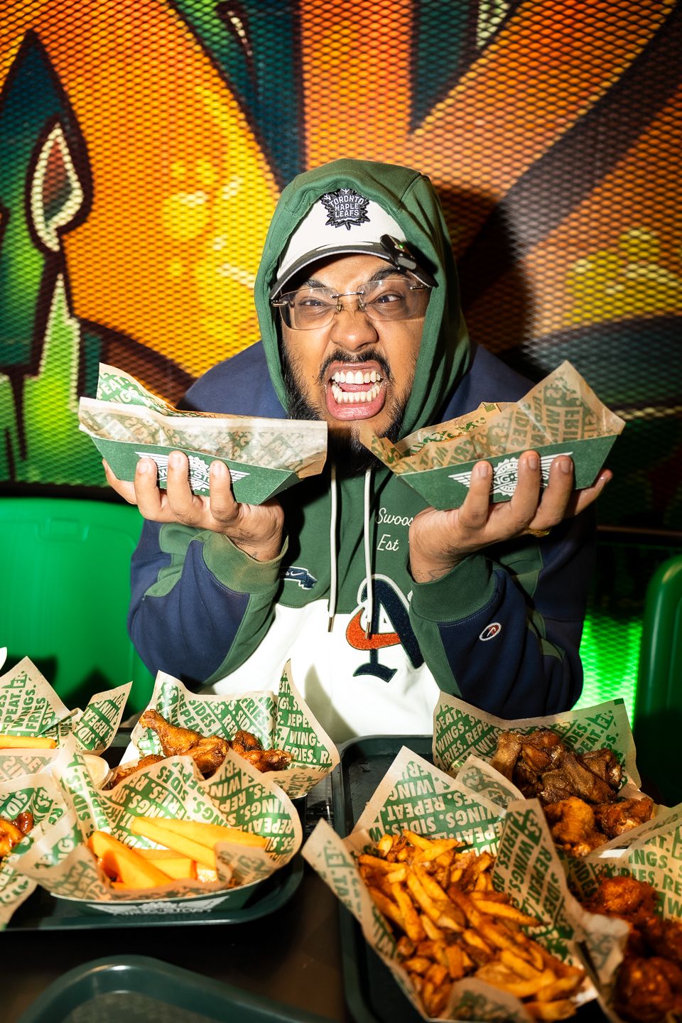 Man in green hoodie excitedly holding chicken wing baskets surrounded by food at Wings.