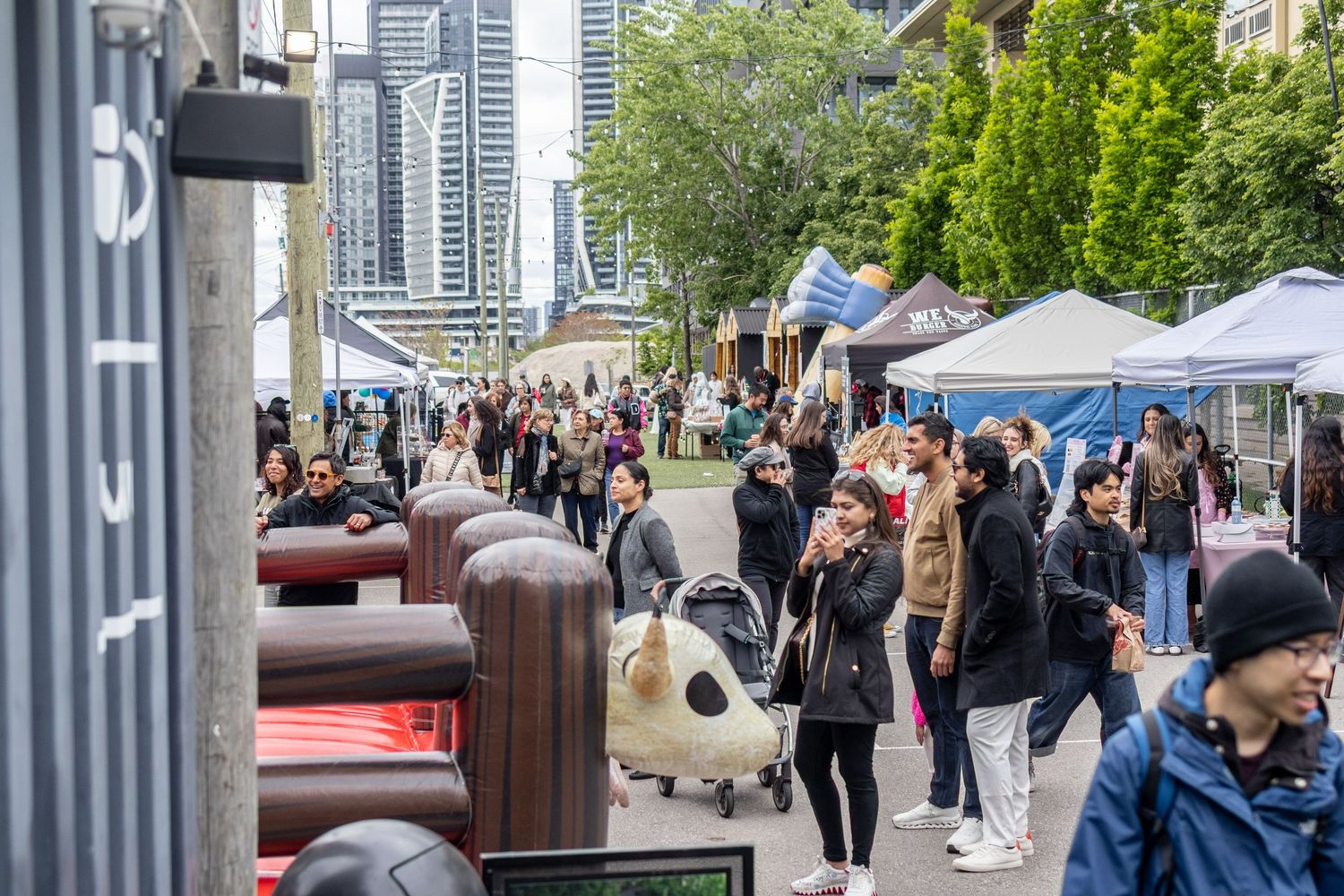 Busy outdoor market with crowds of shoppers walking past vendor tents in an urban setting with skyscrapers.