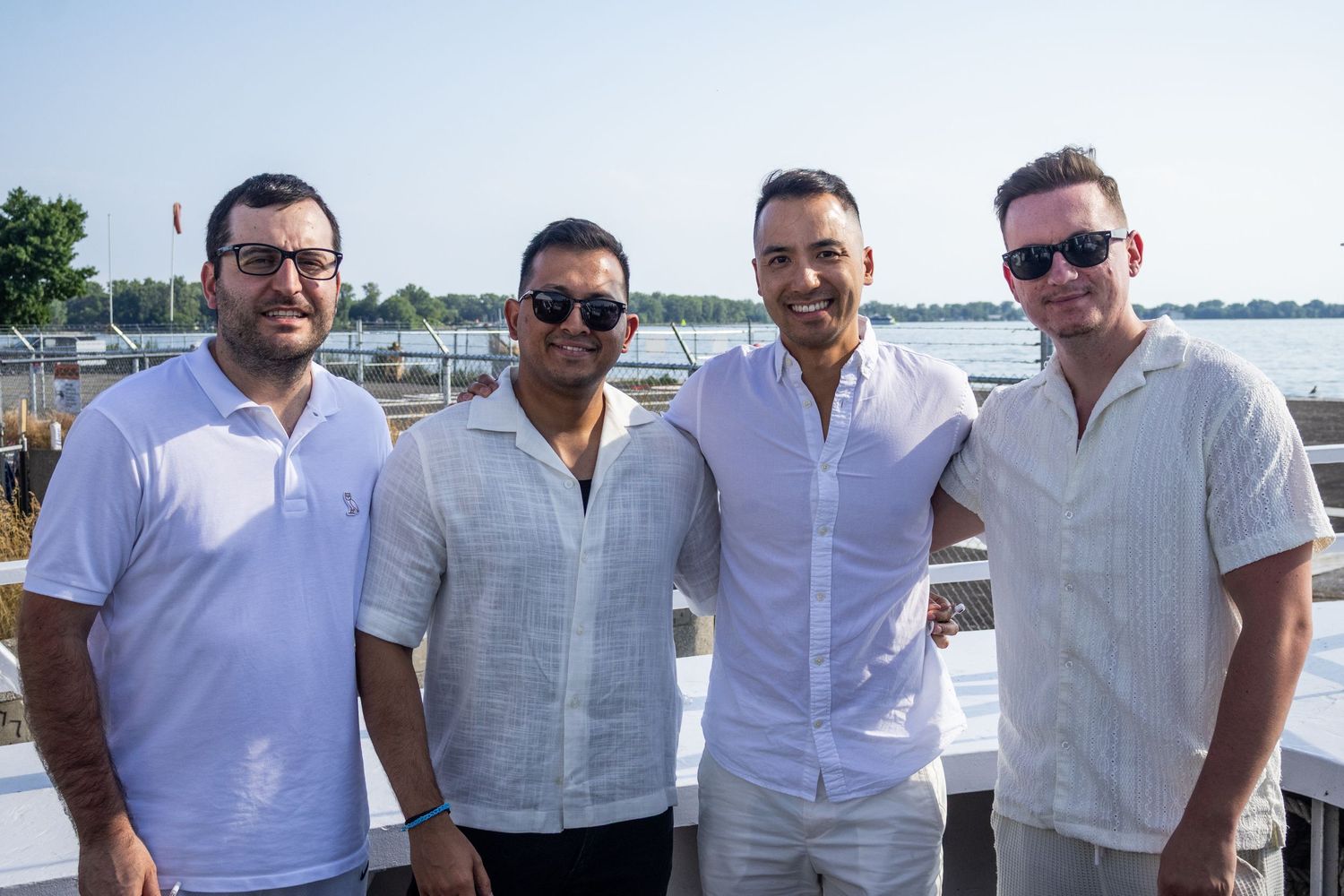 Four men in white shirts smiling together at a waterfront outdoor event.