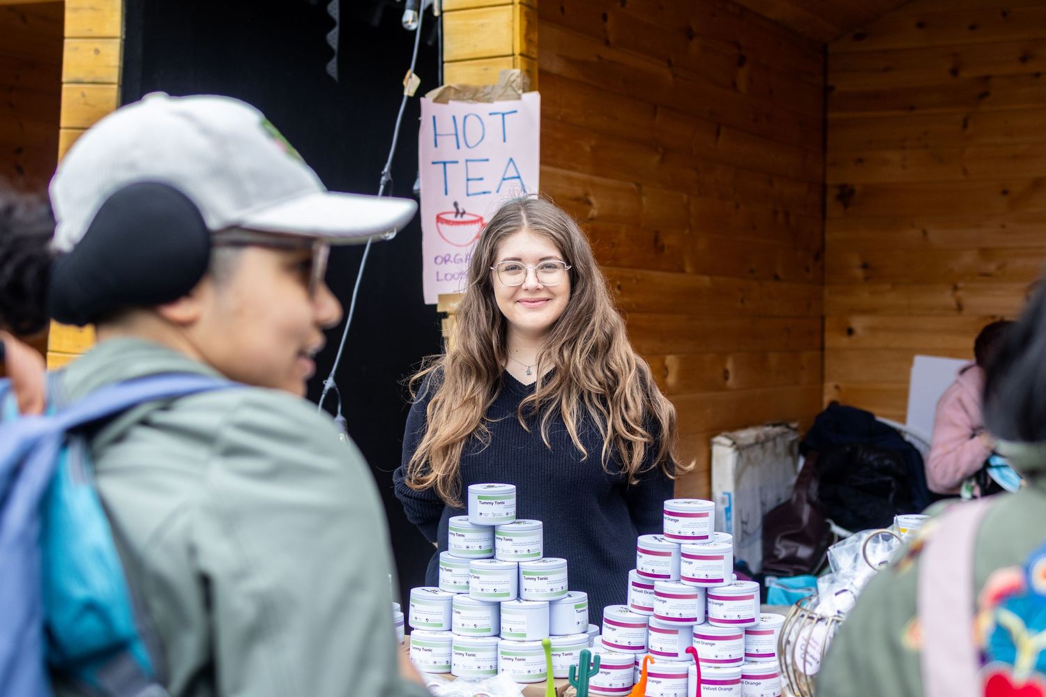 Young woman with long brown hair smiling behind a hot tea stand with stacked cans.