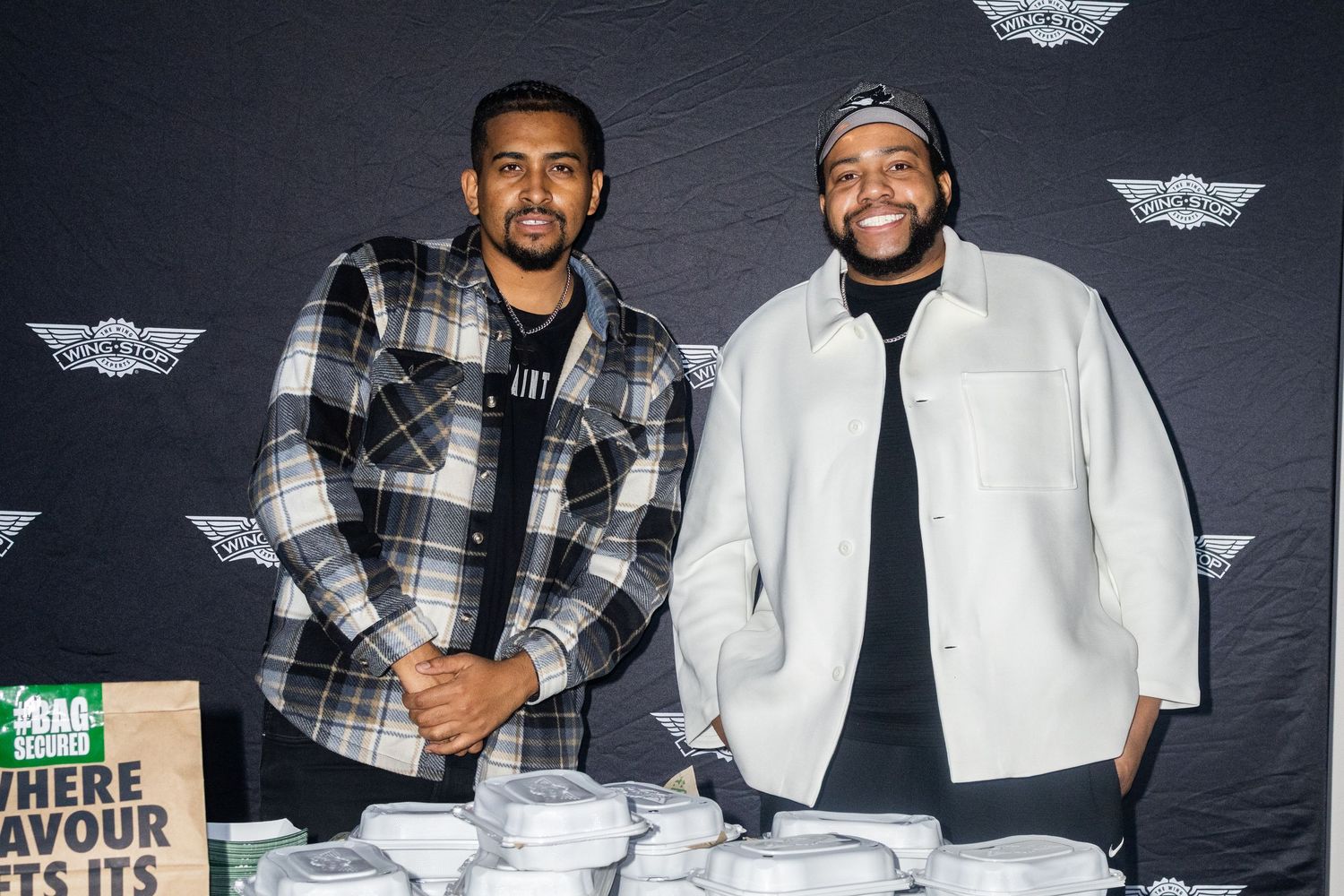 Two men smiling and posing together at a food event in front of a branded backdrop with meal containers.