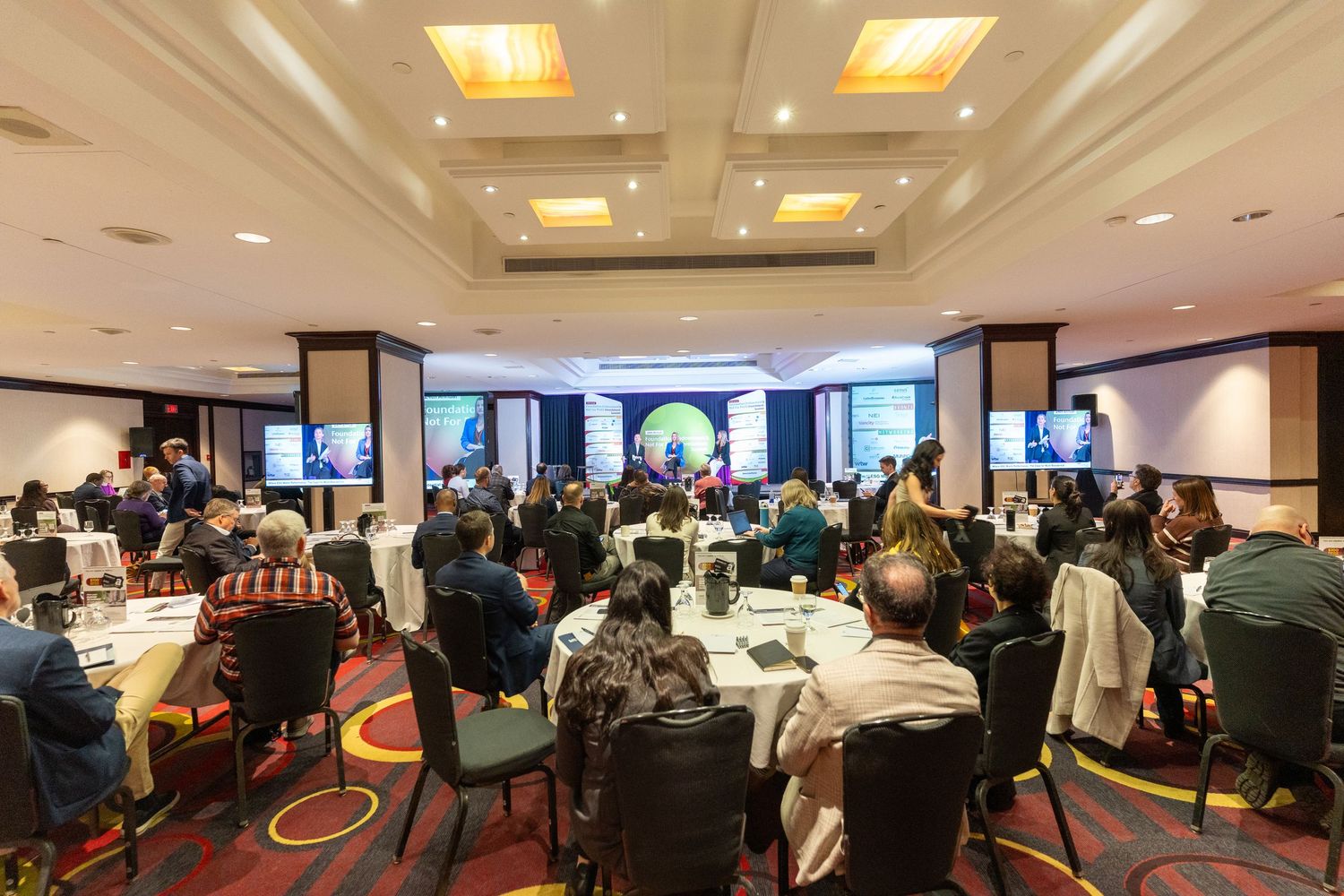 Attendees seated at round tables during a professional conference in a large hotel ballroom.