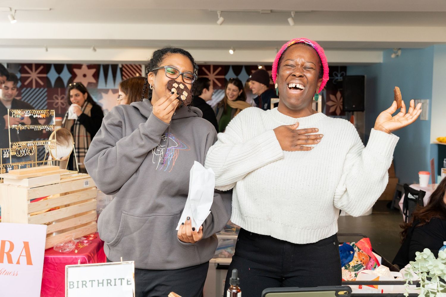 Two joyful women laughing together at a vendor booth inside the Kreate Market Series local market.